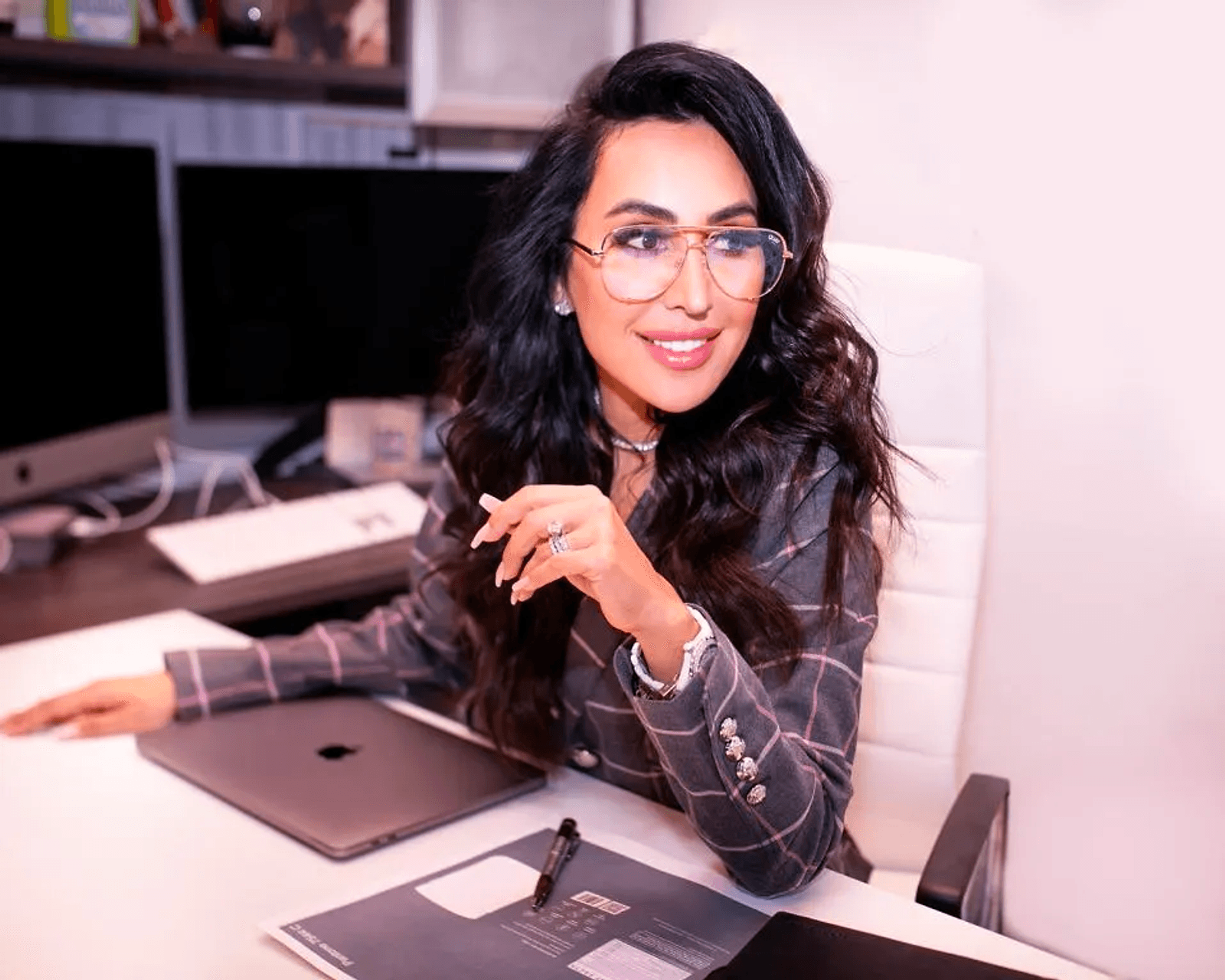 Smiling woman in glasses seated at a desk with a closed laptop, desktop monitor, and papers.