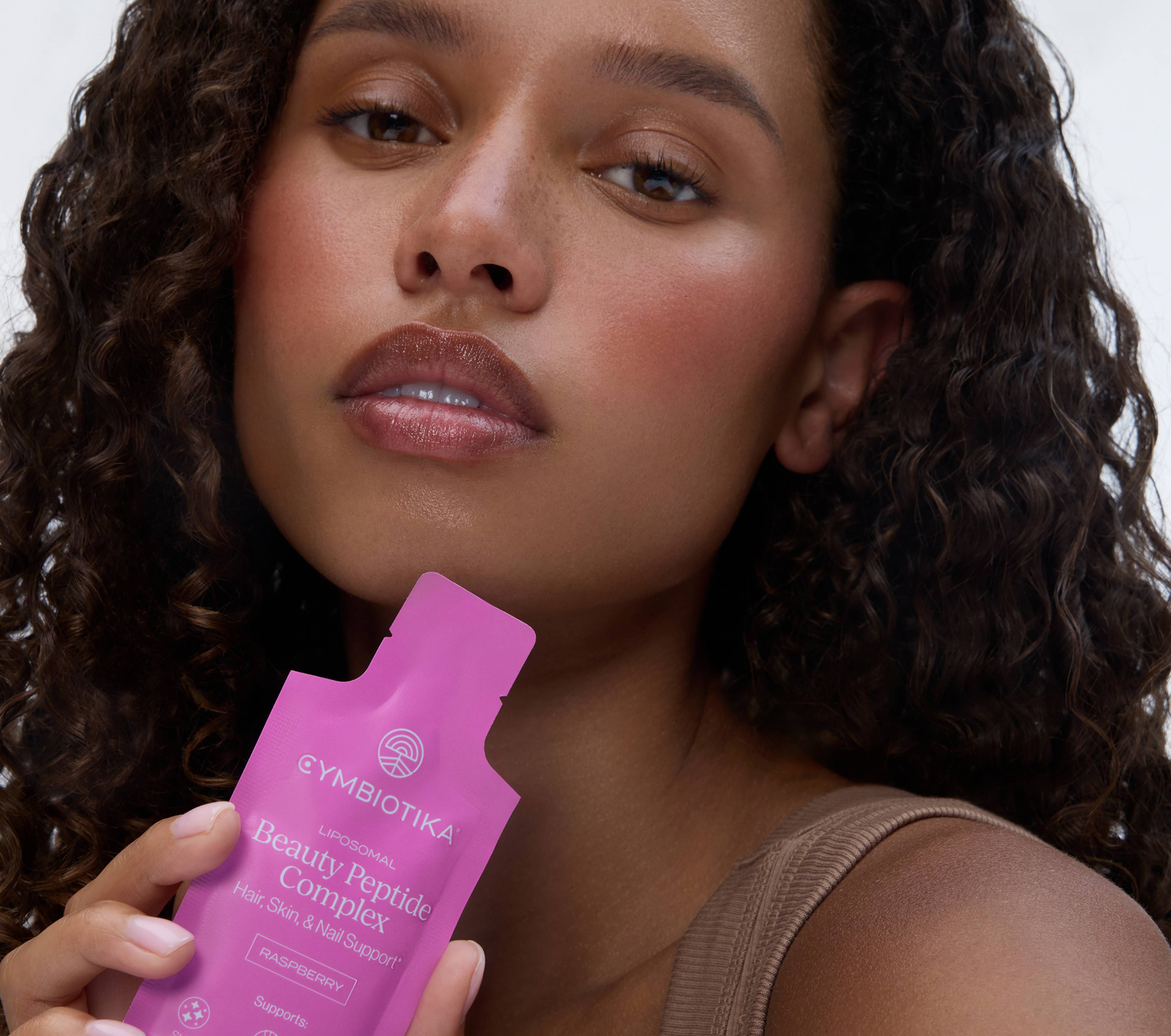 Woman holding a pink Cymbiotika Liposomal Beauty Peptide Complex sachet near her face.