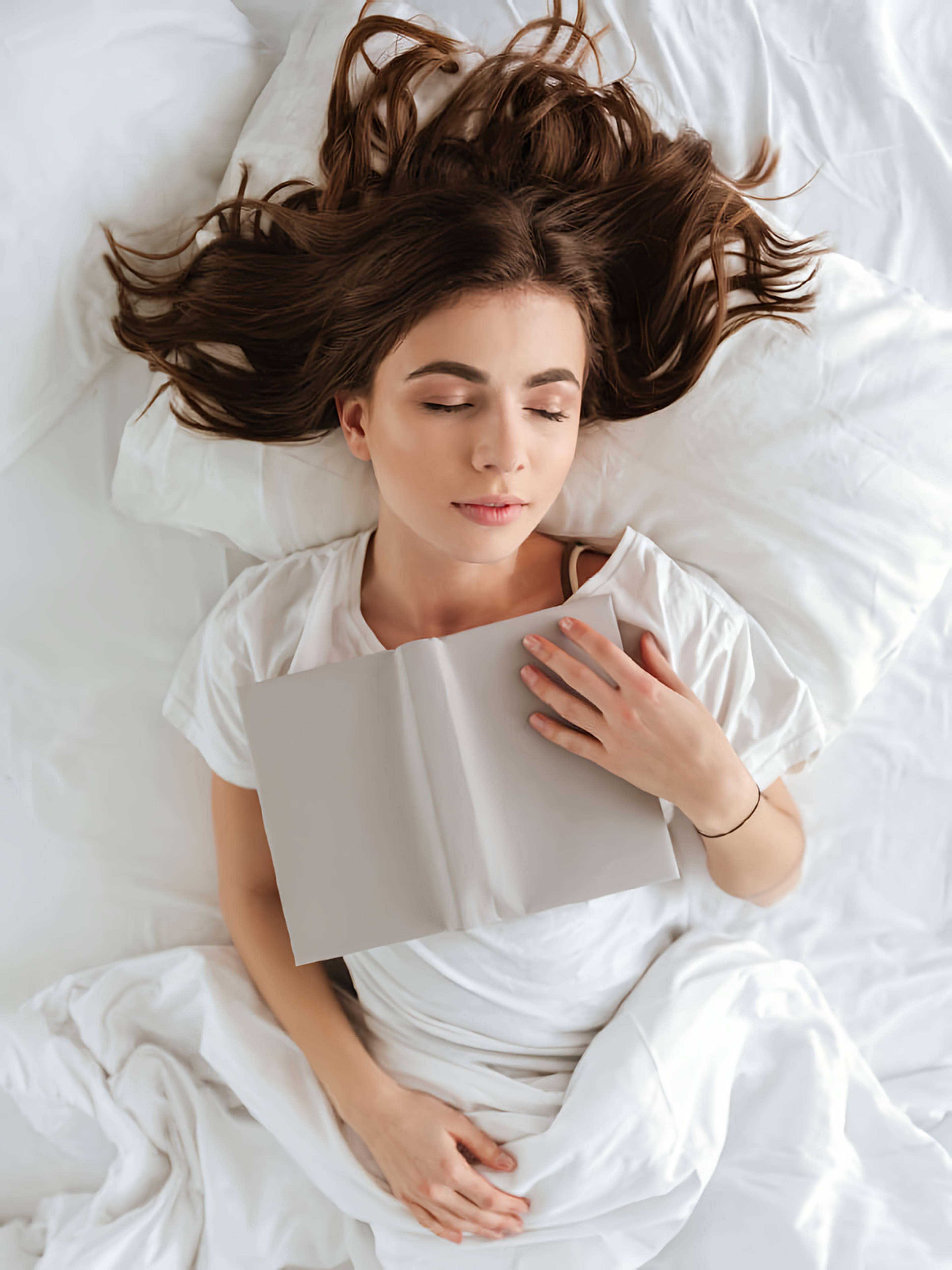 Woman resting on white bedding with an open book on her chest