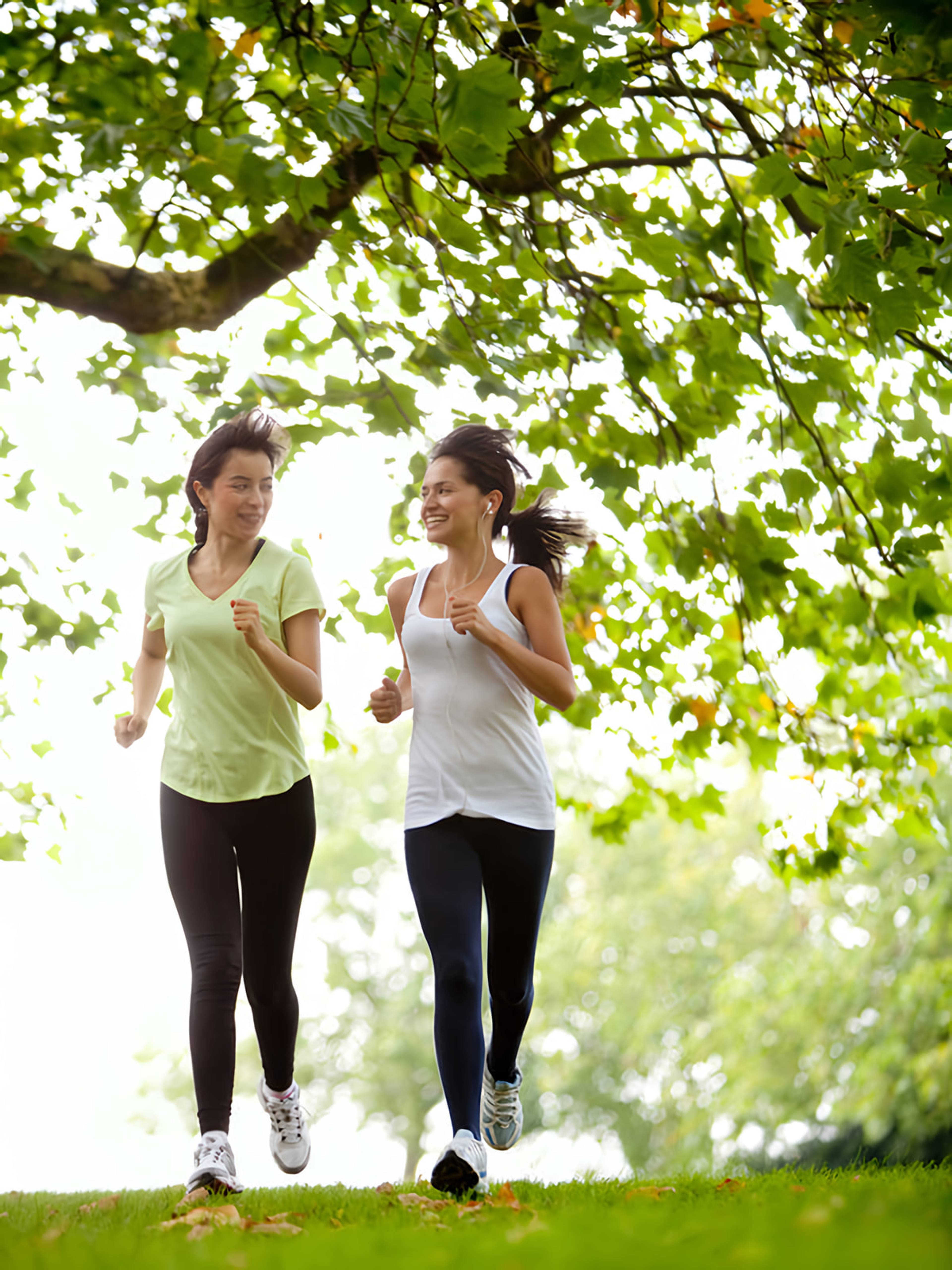 Two women jogging together beneath leafy trees in a park.