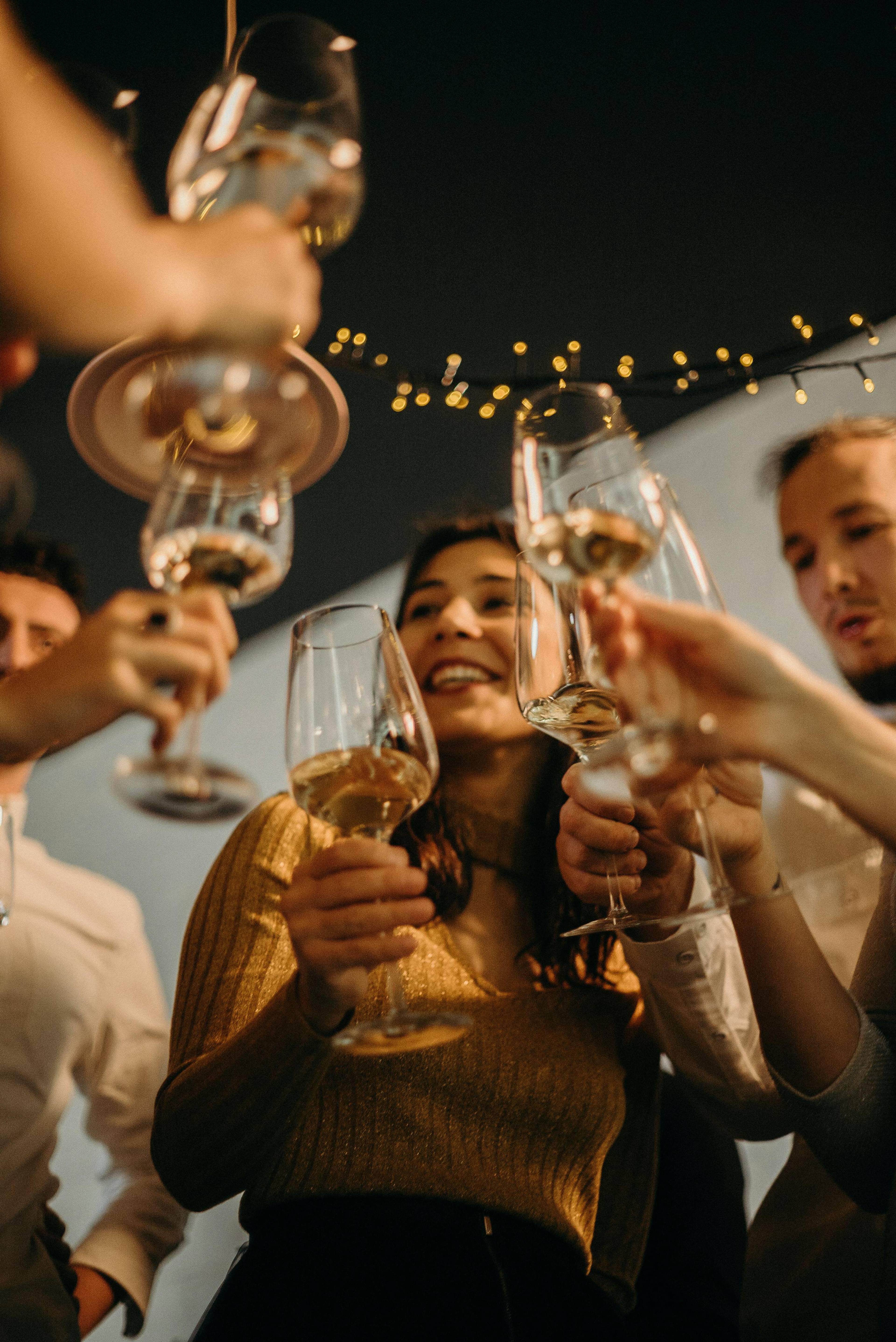 People raising stemmed glasses together beneath string lights.