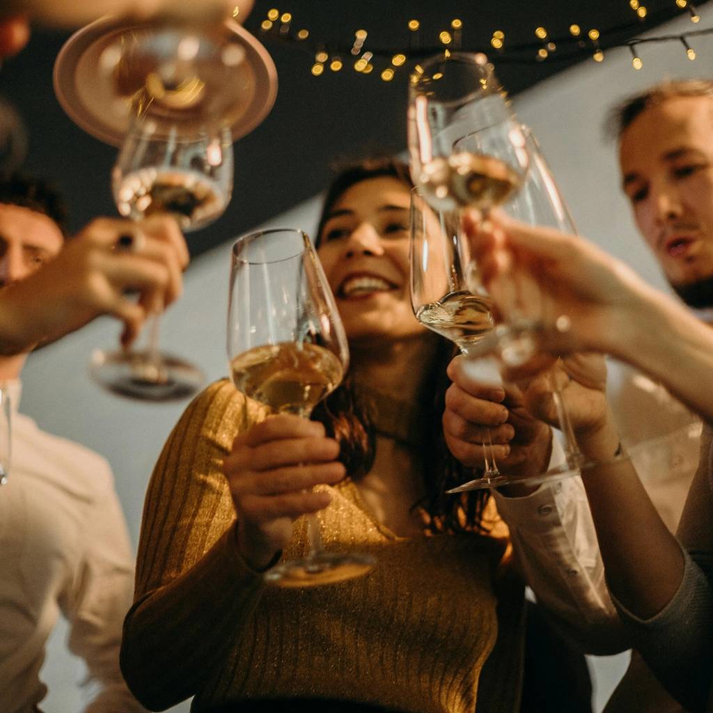 People raising stemmed glasses together beneath string lights.