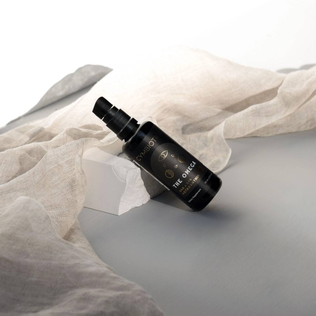 Black pump bottle labeled THE OMEGA leans against a white block, surrounded by beige draped fabric.