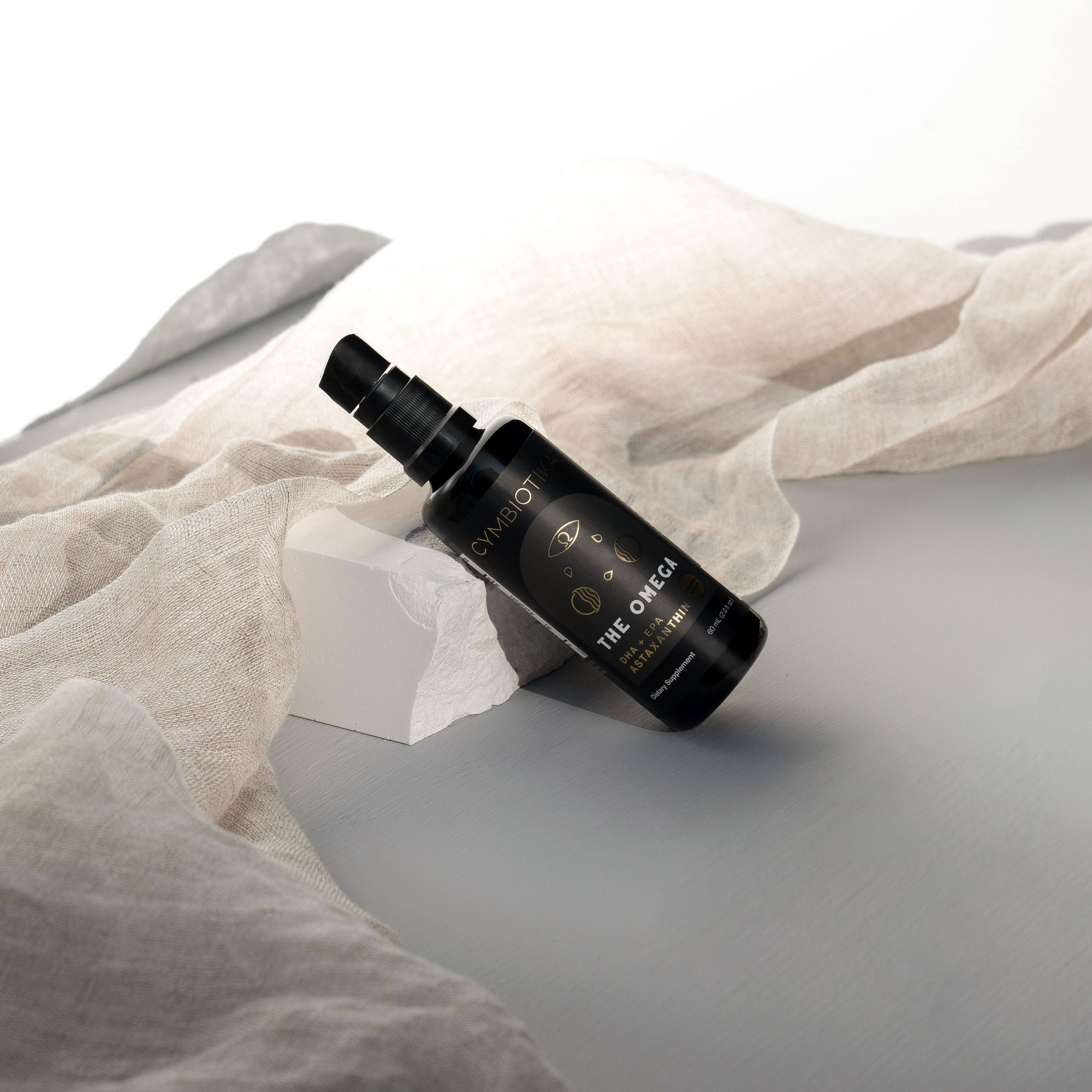 Black pump bottle labeled THE OMEGA leans against a white block, surrounded by beige draped fabric.
