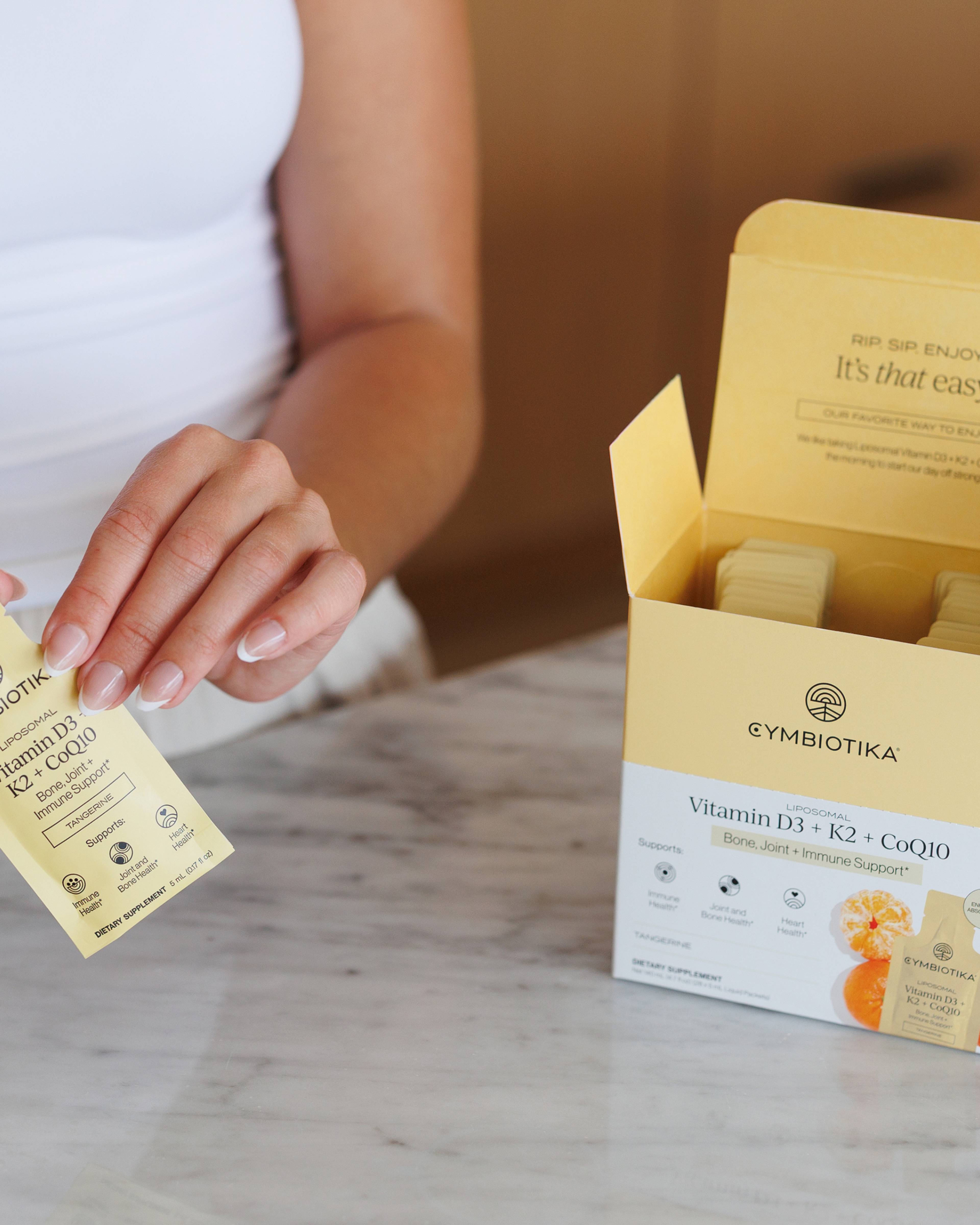 A person holds a Vitamin D3 + K2 + CoQ10 sachet beside its open yellow-and-white Cymbiotika box.