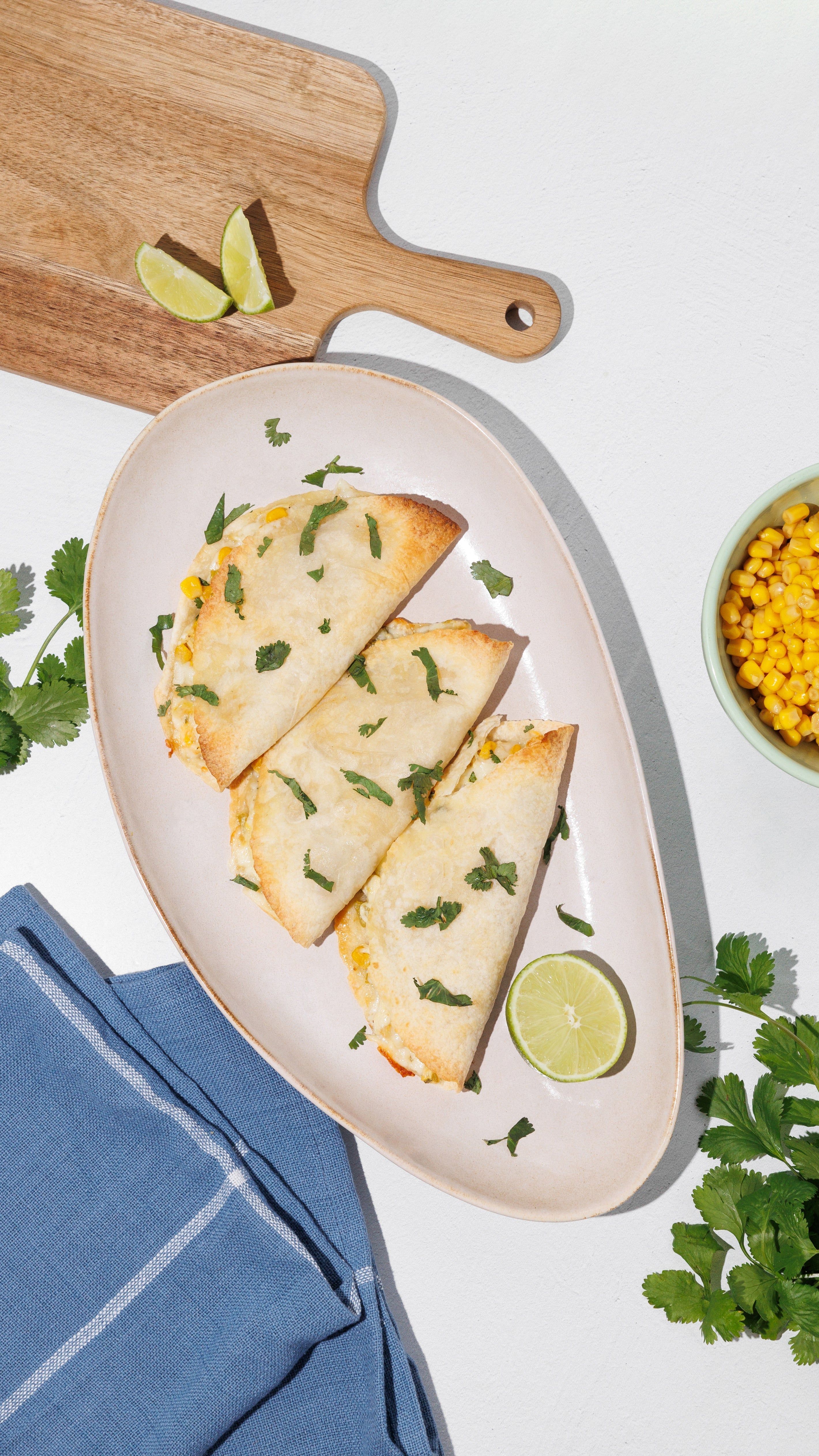 Three folded, lightly browned tortillas on an oval plate with lime, cilantro, corn, and a blue napkin.