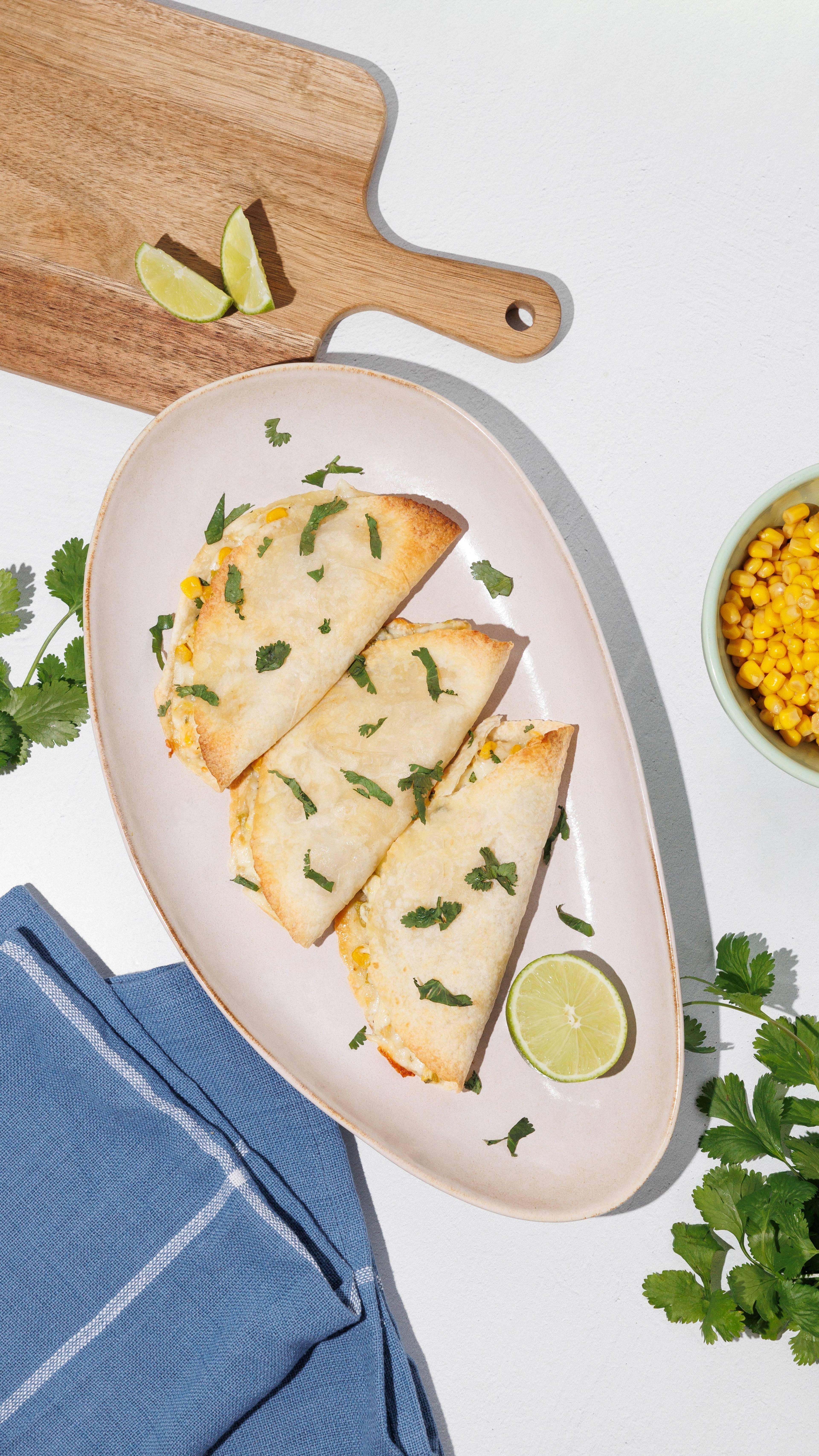 Three folded, lightly browned tortillas on an oval plate with lime, cilantro, corn, and a blue napkin.