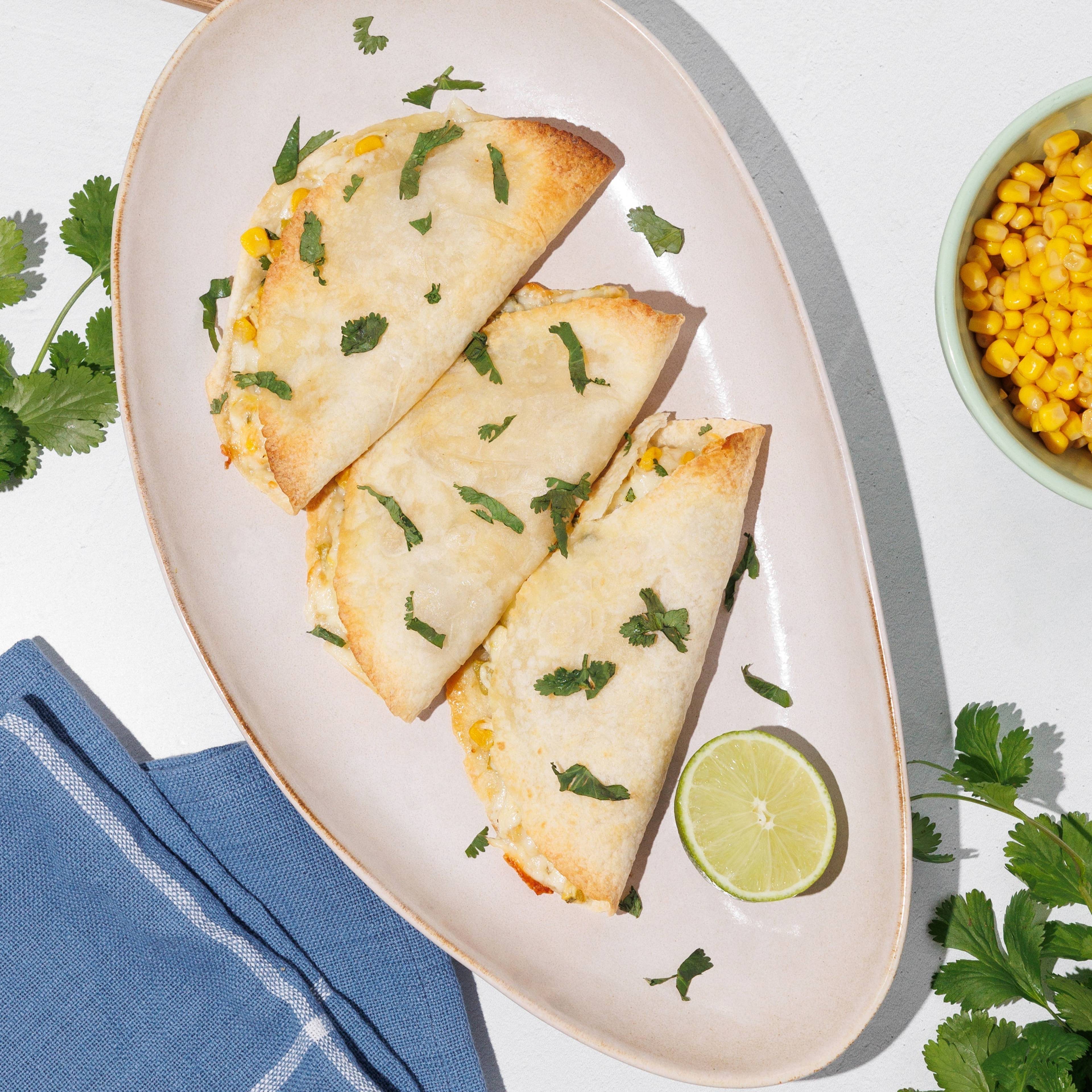 Three folded, lightly browned tortillas on an oval plate with lime, cilantro, corn, and a blue napkin.
