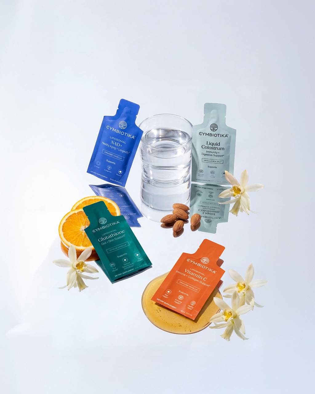 Cymbiotika NAD+, Liquid Colostrum, Glutathione, and Vitamin C sachets arranged with water, orange, almonds, and flowers.