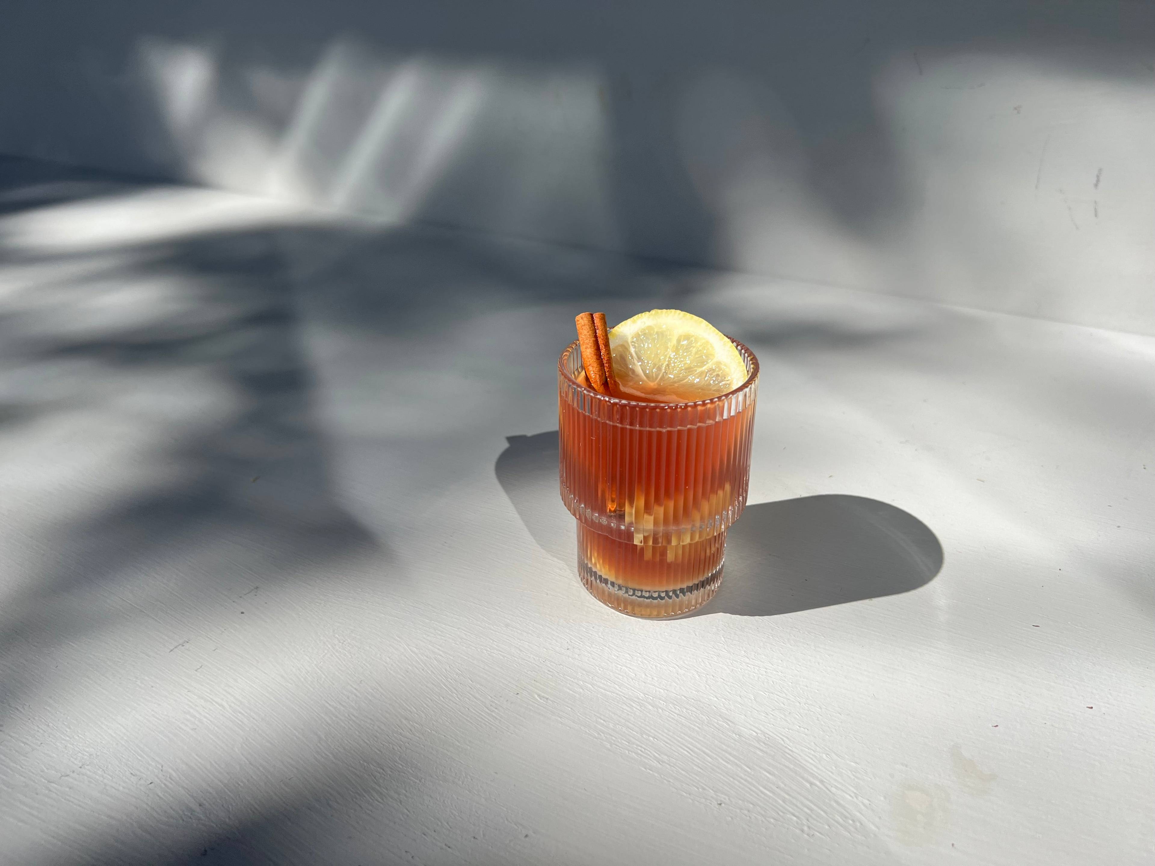 Ribbed glass of amber drink garnished with a lemon slice and cinnamon stick on a sunlit white table.