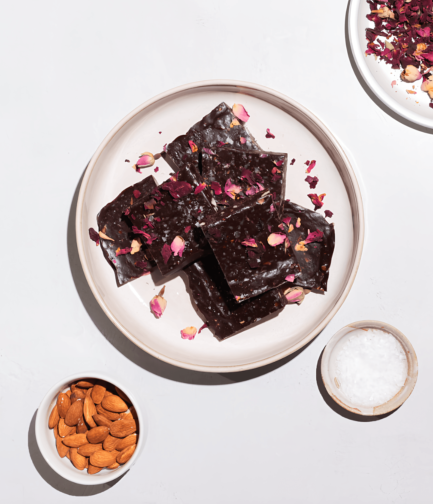 Dark chocolate bark squares topped with rose petals on a plate, with almonds and salt nearby.