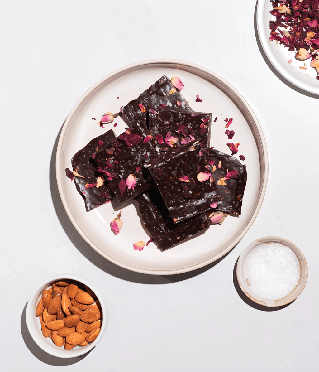 Dark chocolate bark squares topped with rose petals on a plate, with almonds and salt nearby.
