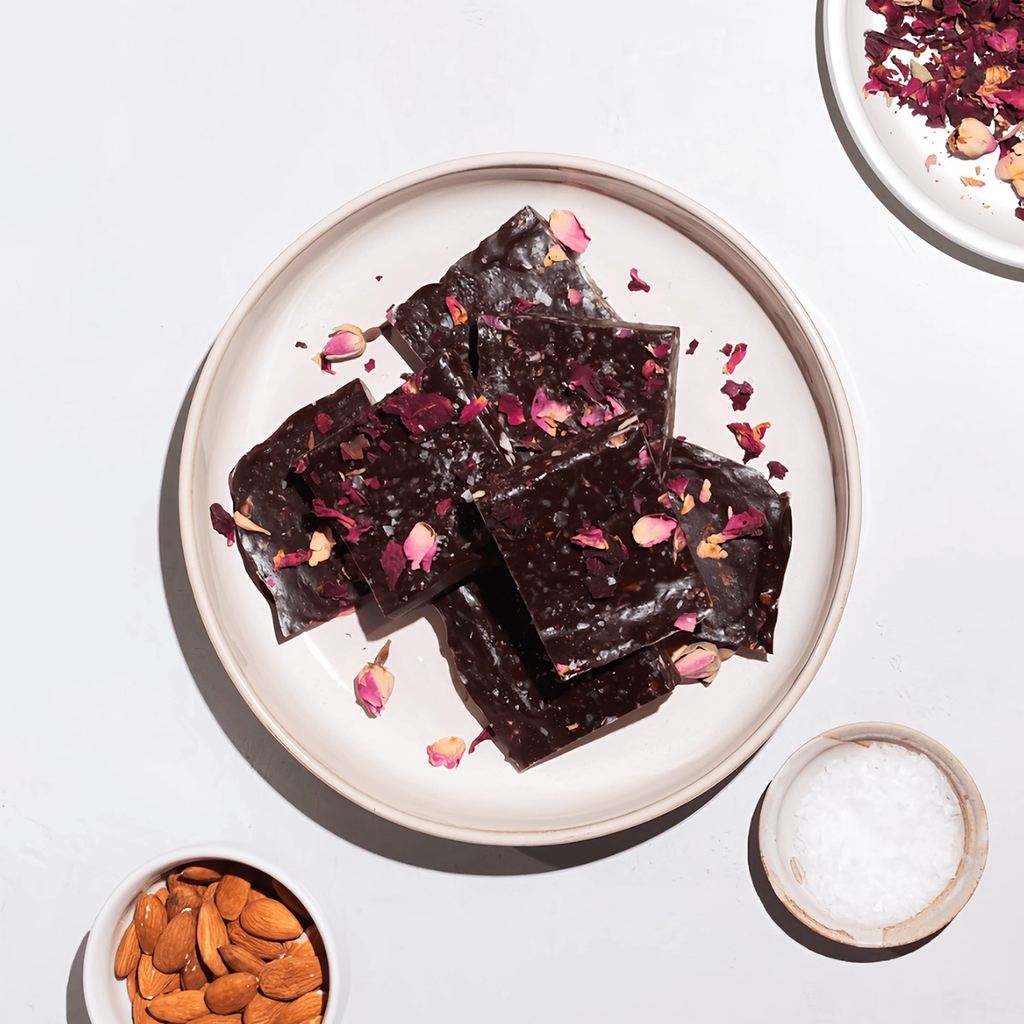 Dark chocolate bark squares topped with rose petals on a plate, with almonds and salt nearby.