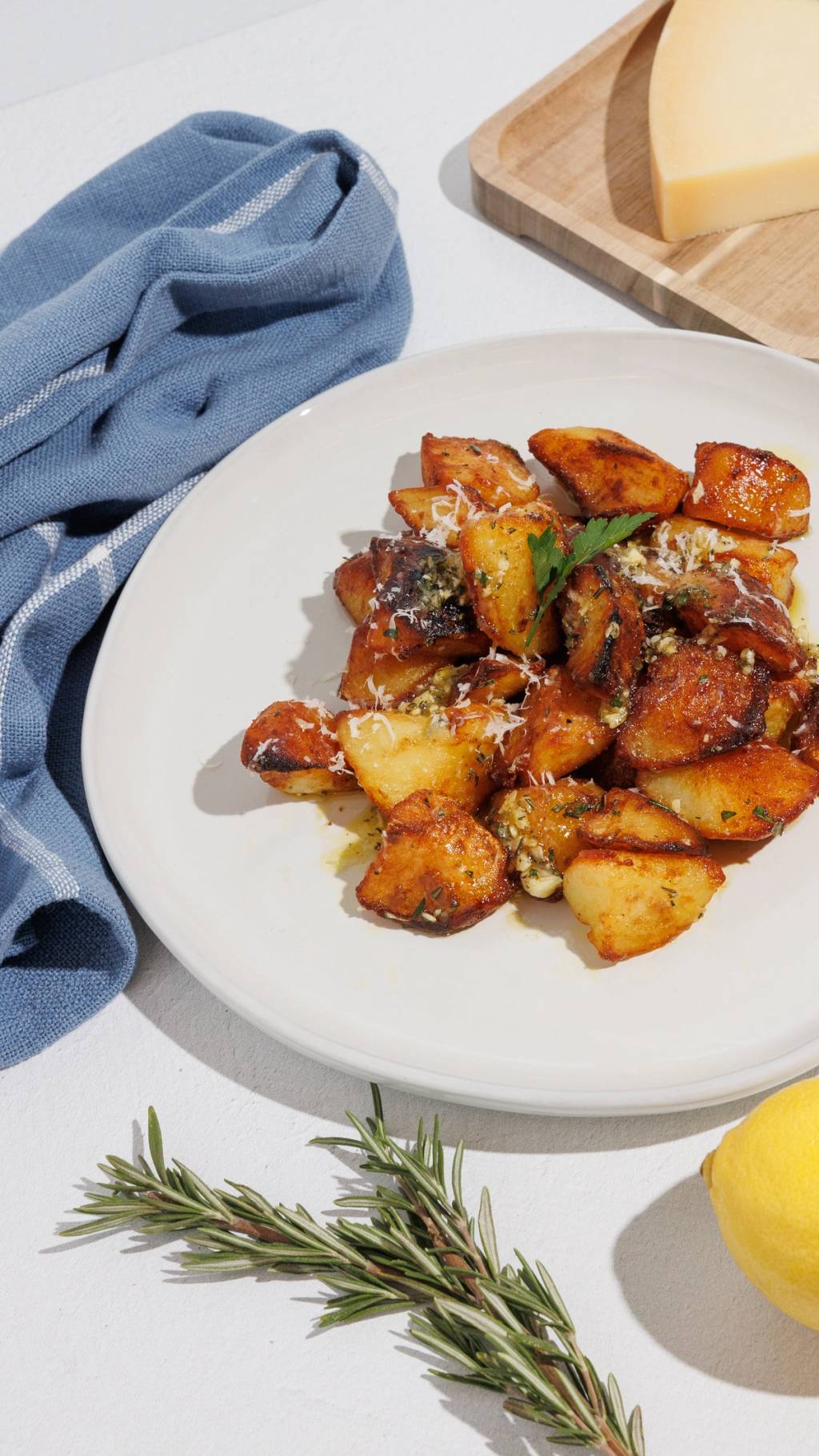Plate of crispy herb-roasted potato bites with grated cheese, rosemary, lemon, and a wedge of cheese nearby.