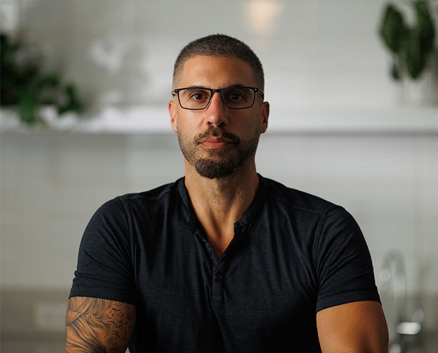 Man wearing glasses and a dark shirt seated indoors with plants in the background.