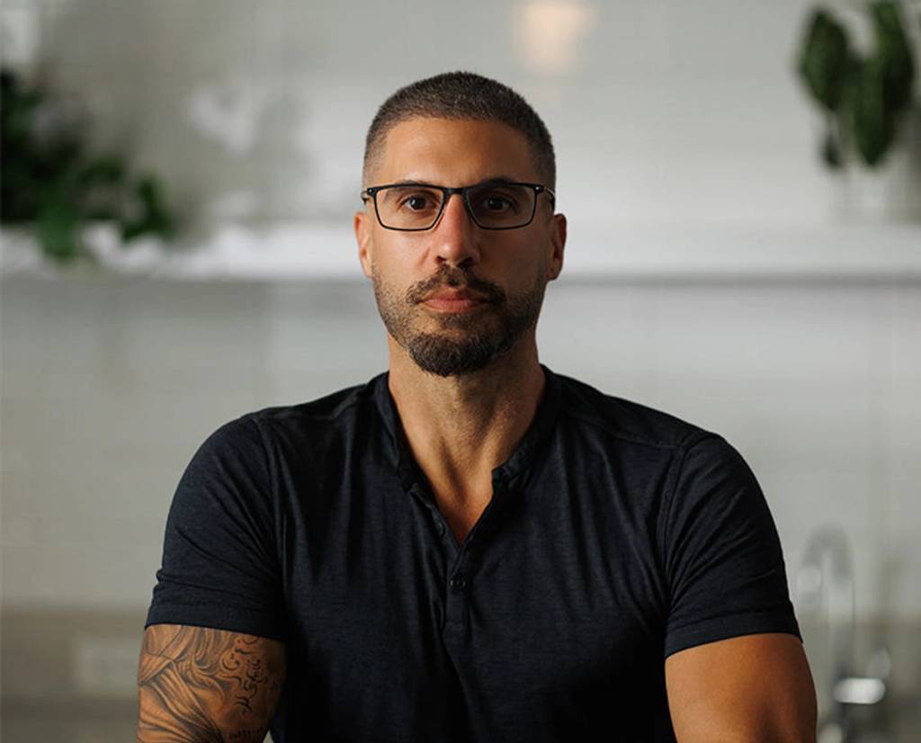 Man wearing glasses and a dark shirt seated indoors with plants in the background.
