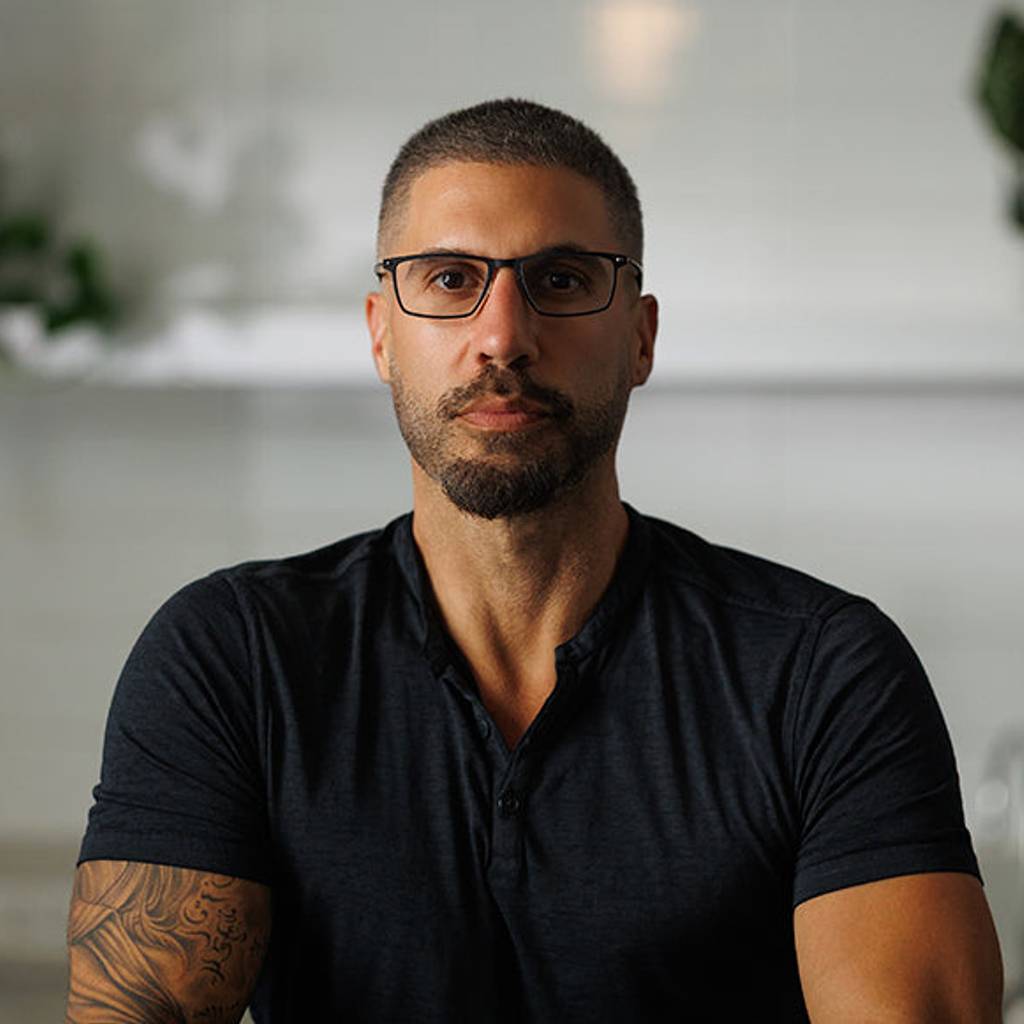 Man wearing glasses and a dark shirt seated indoors with plants in the background.
