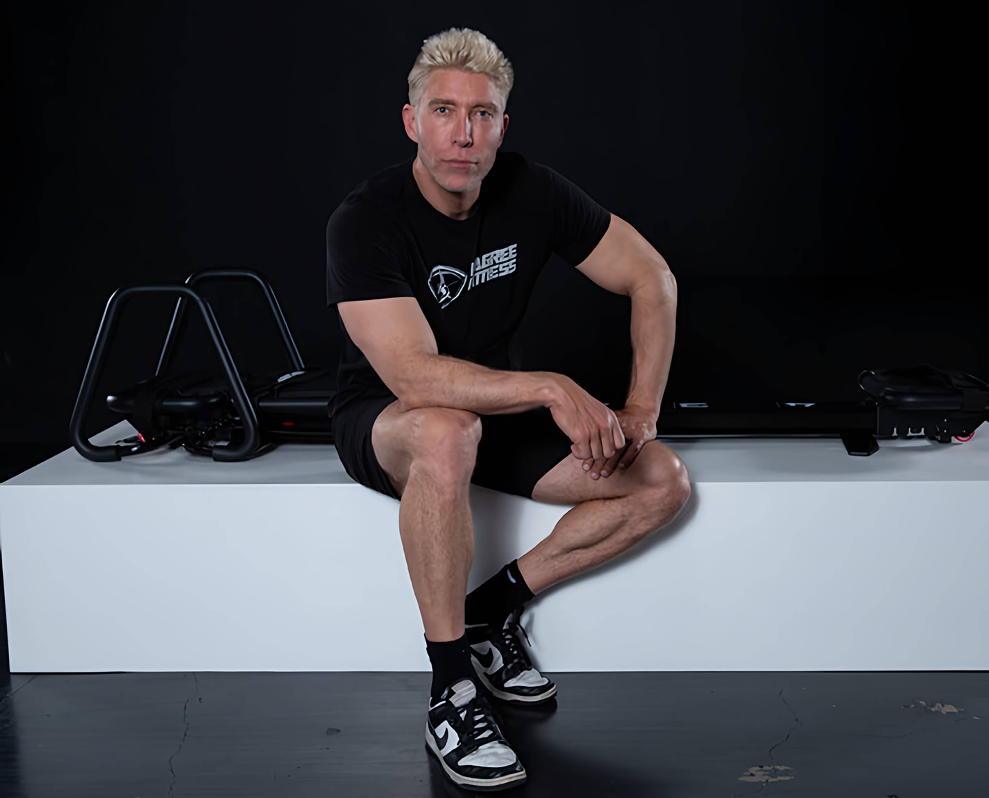 Man in black Lagree Fitness clothing sits on a white exercise platform, with workout machines behind him.