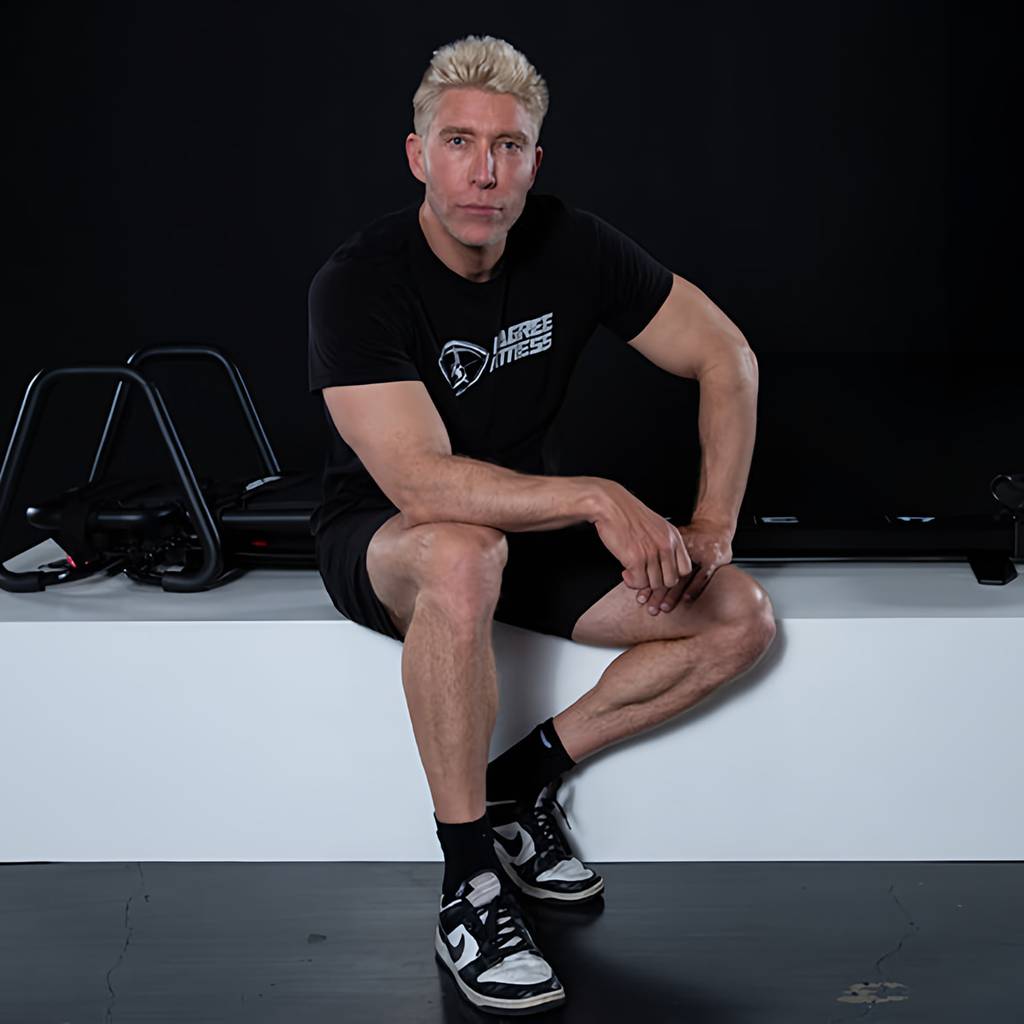 Man in black Lagree Fitness clothing sits on a white exercise platform, with workout machines behind him.