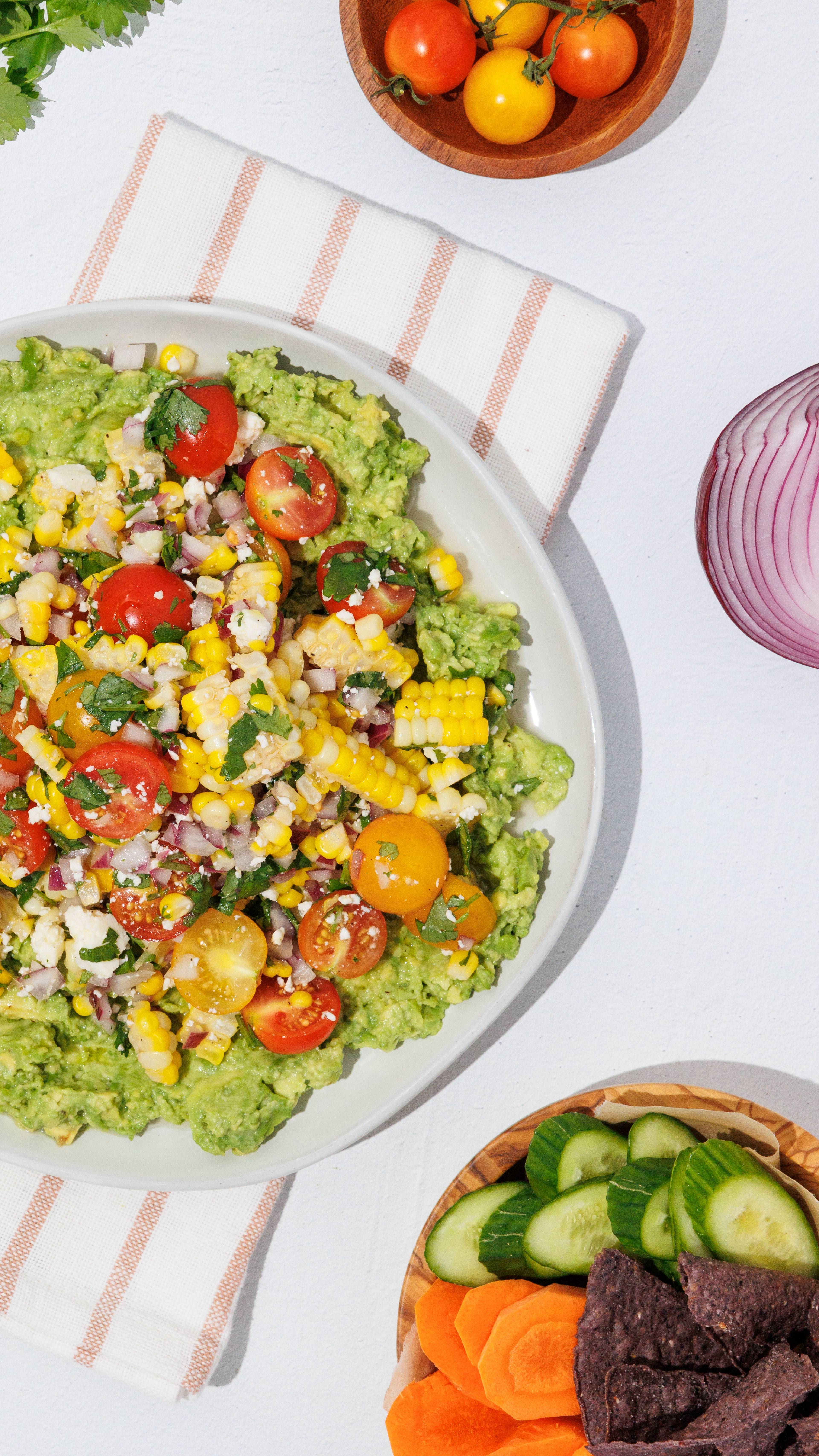 Smashed avocado topped with corn salsa, cherry tomatoes, and herbs, served with cucumber, carrots, and tortilla chips.