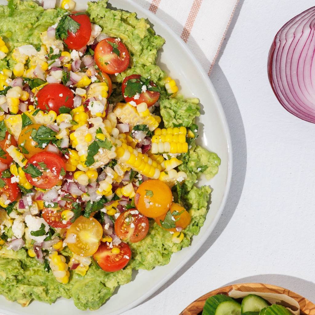 Smashed avocado topped with corn salsa, cherry tomatoes, and herbs, served with cucumber, carrots, and tortilla chips.