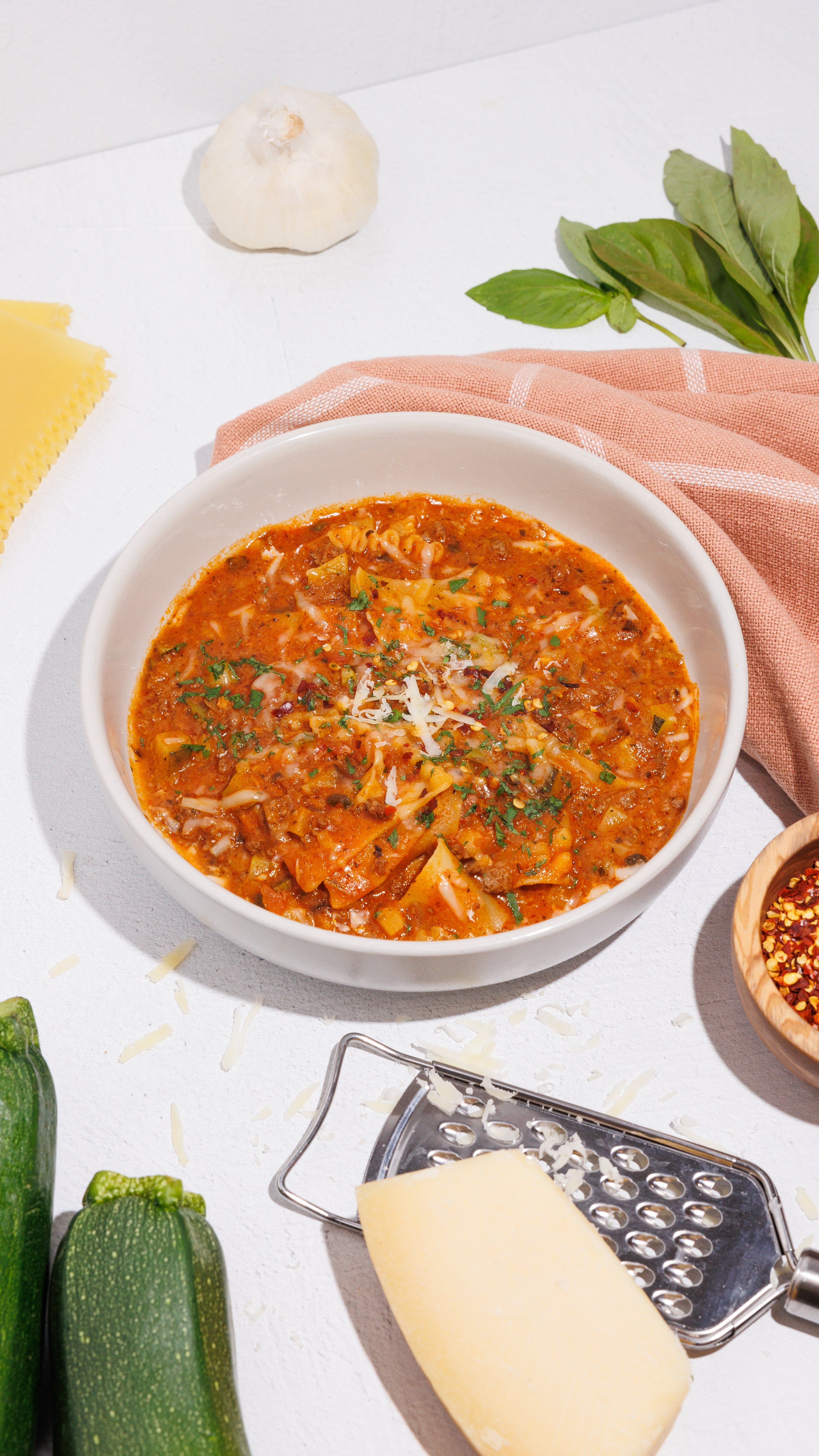 Bowl of lasagna soup topped with herbs and grated cheese, surrounded by garlic, basil, zucchini, pasta, and cheese.