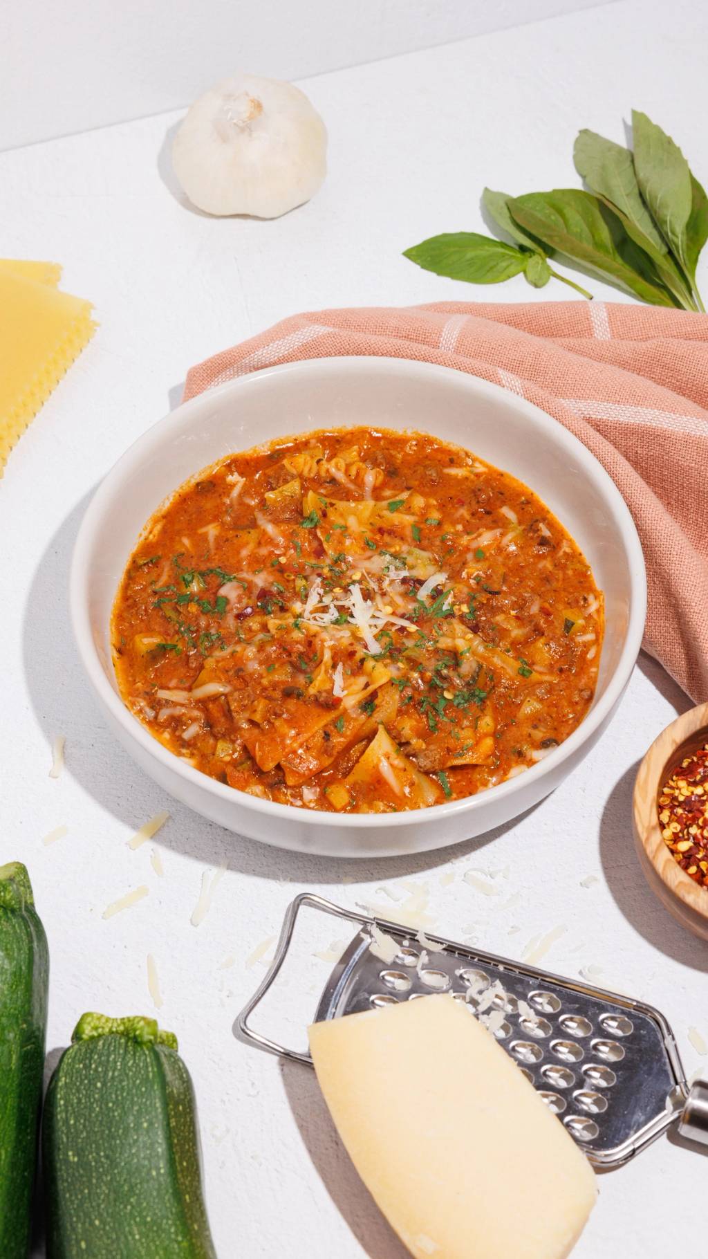 Bowl of lasagna soup topped with herbs and grated cheese, surrounded by garlic, basil, zucchini, pasta, and cheese.