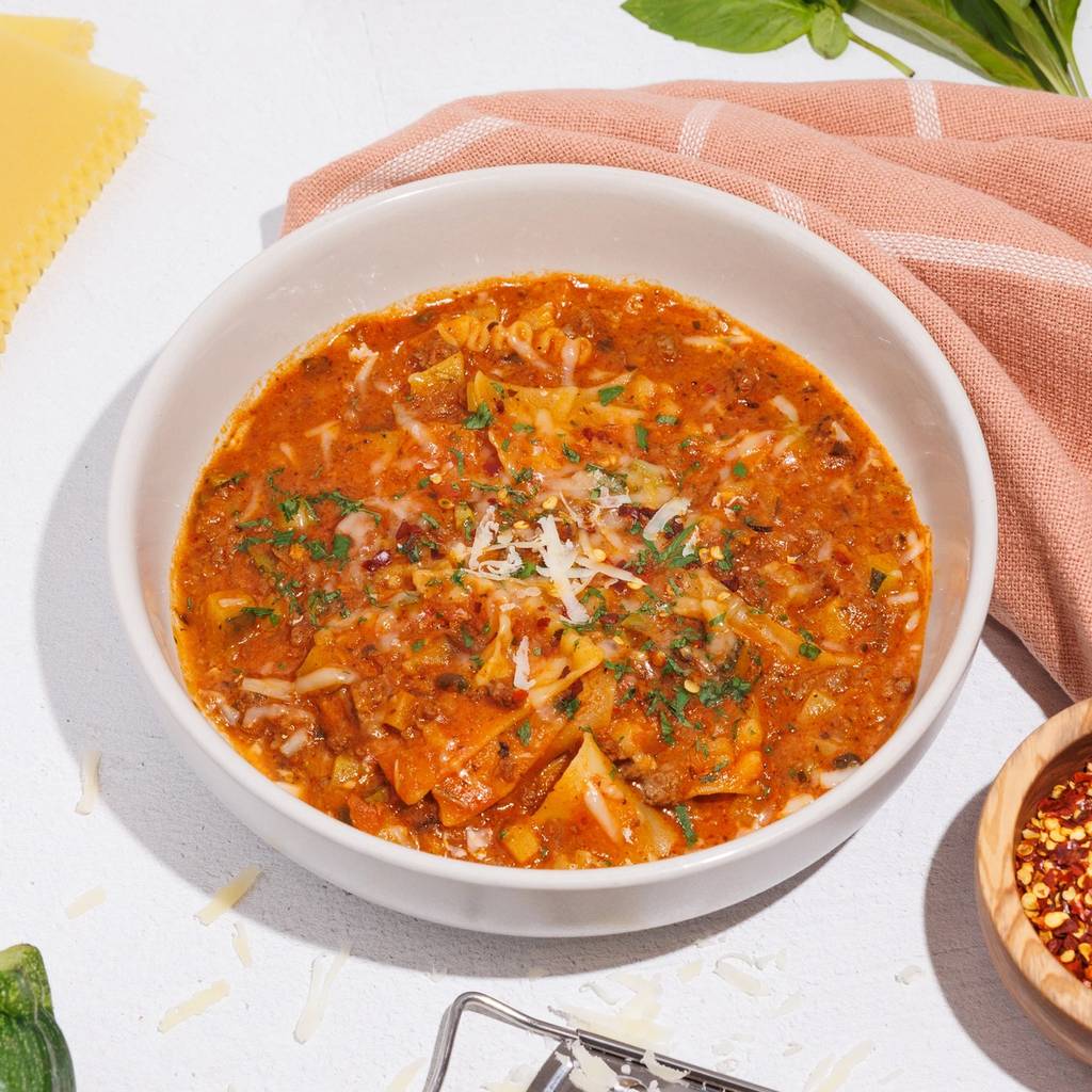 Bowl of lasagna soup topped with herbs and grated cheese, surrounded by garlic, basil, zucchini, pasta, and cheese.
