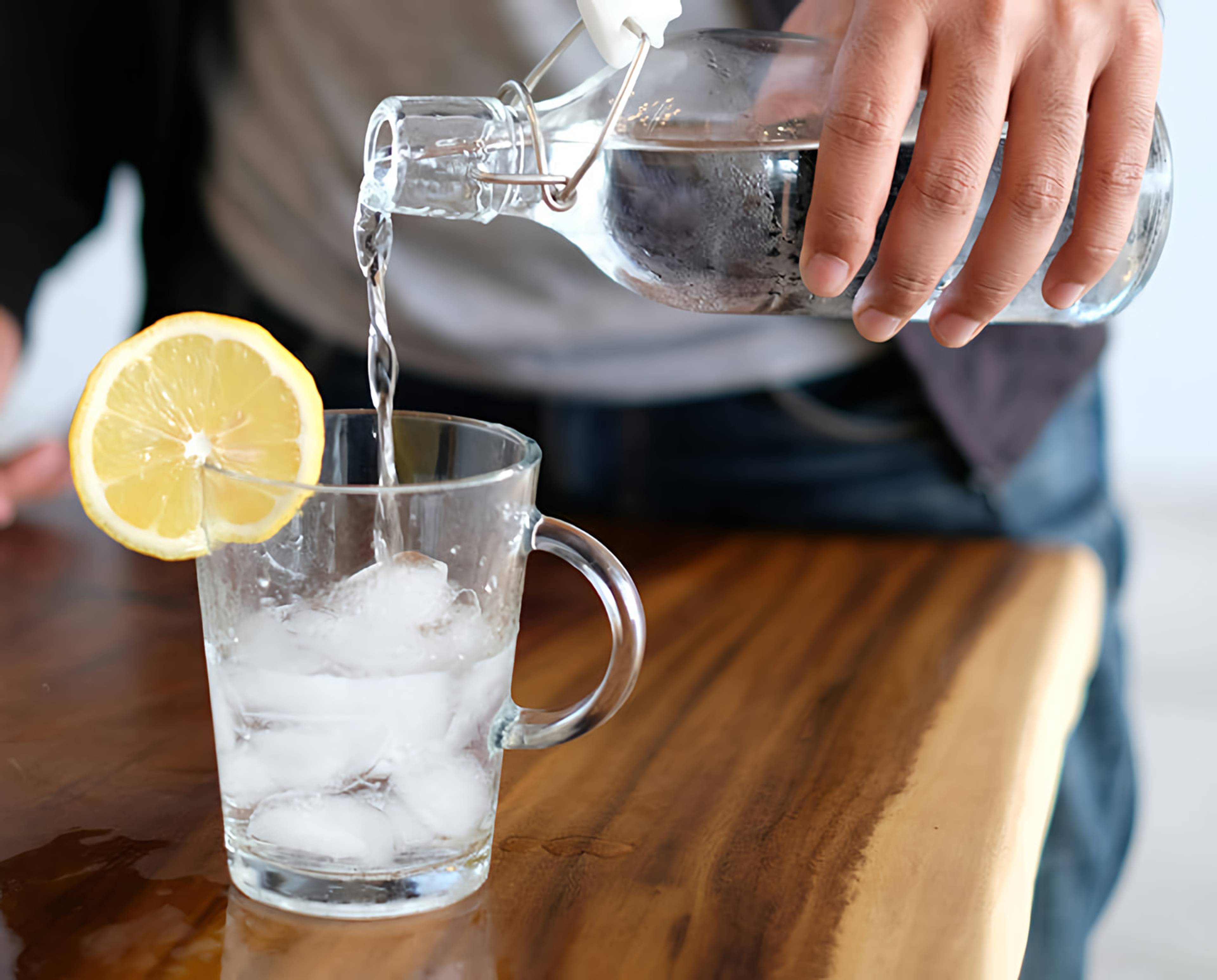 Person pouring water from a glass bottle into an ice-filled glass with a lemon slice.