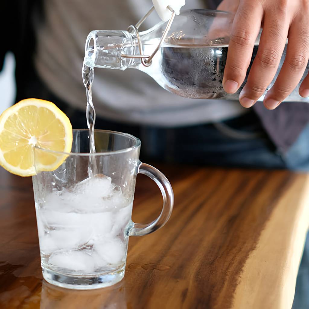 Person pouring water from a glass bottle into an ice-filled glass with a lemon slice.