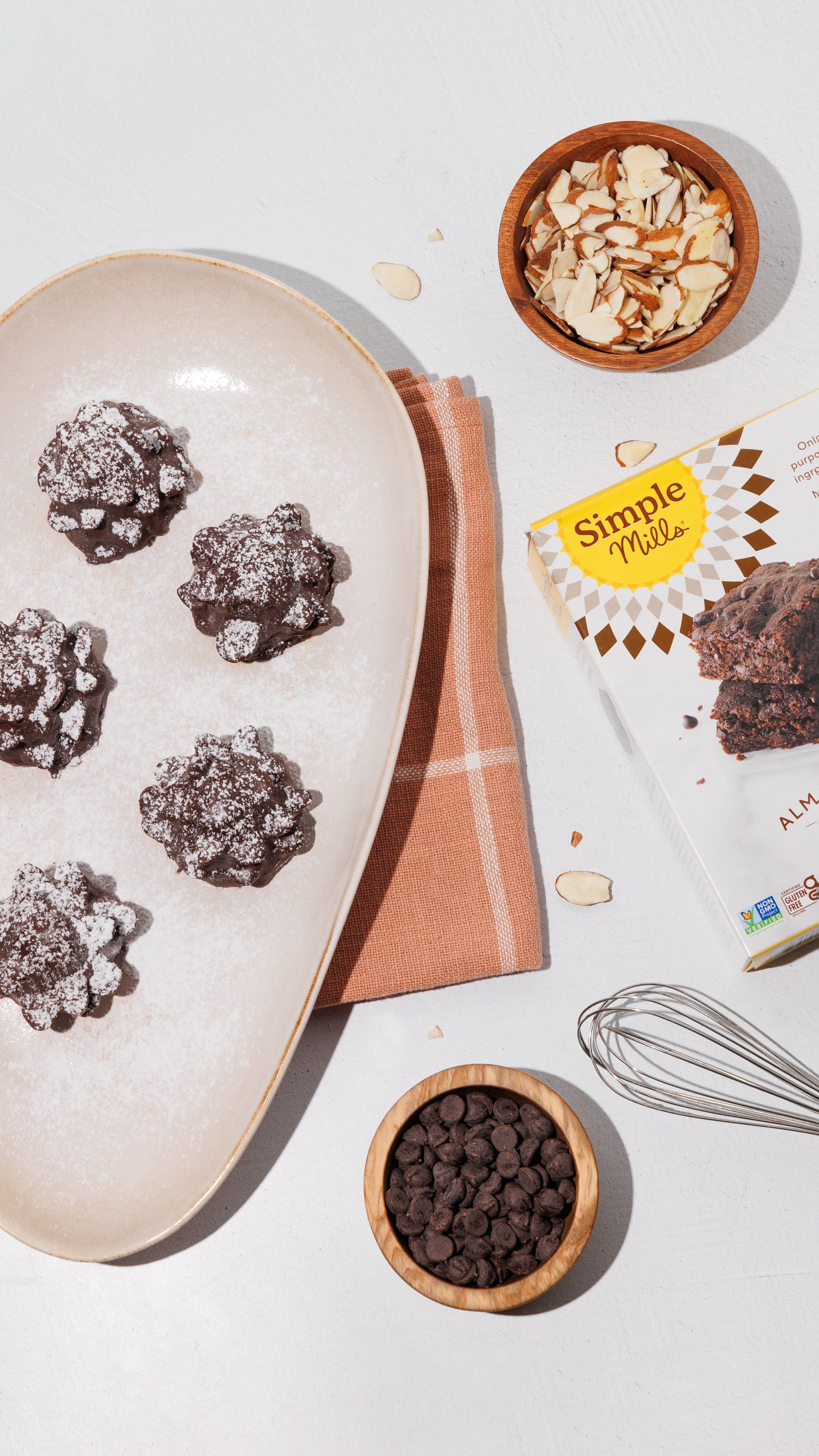 Overhead view of powdered sugar-dusted chocolate brownies with almonds, chocolate chips, a whisk, and baking mix.