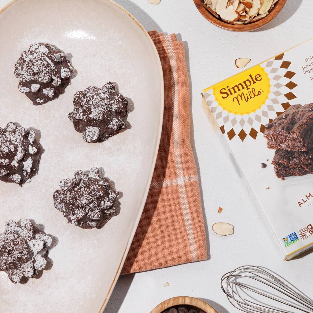 Overhead view of powdered sugar-dusted chocolate brownies with almonds, chocolate chips, a whisk, and baking mix.