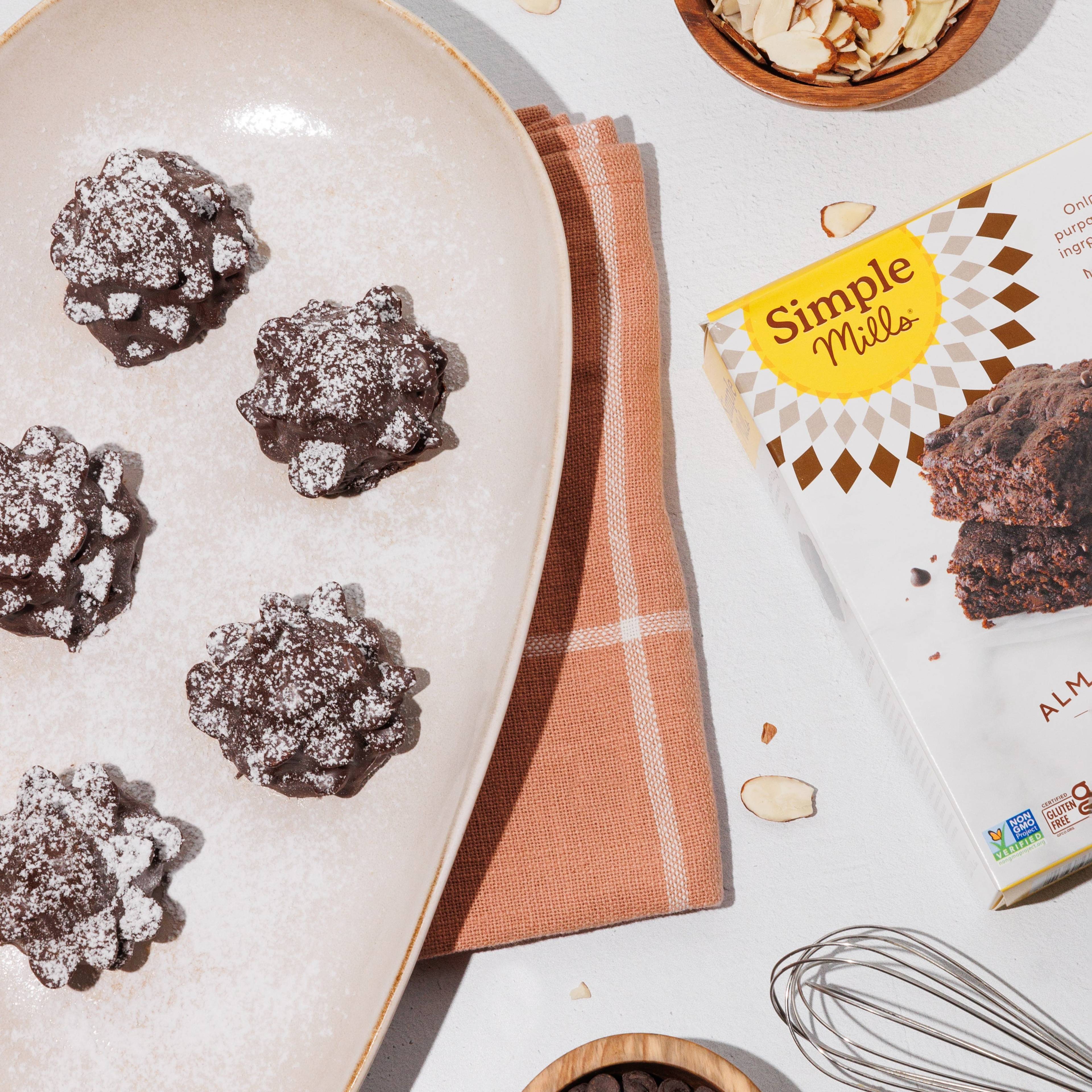 Overhead view of powdered sugar-dusted chocolate brownies with almonds, chocolate chips, a whisk, and baking mix.