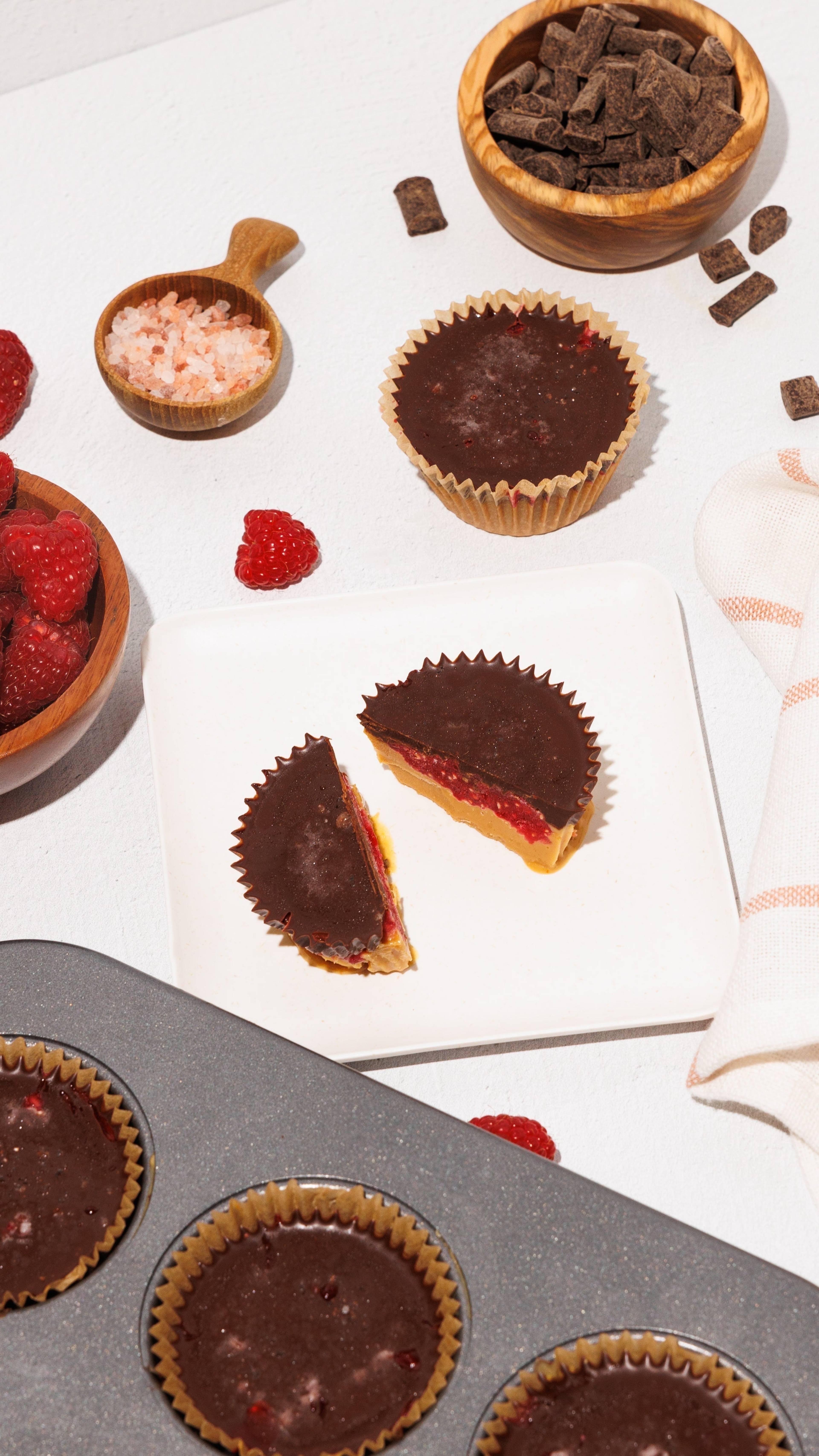 Chocolate-topped cups, including a cut-open cup with red filling and a yellow base, alongside raspberries, chocolate chunks, and pink salt.