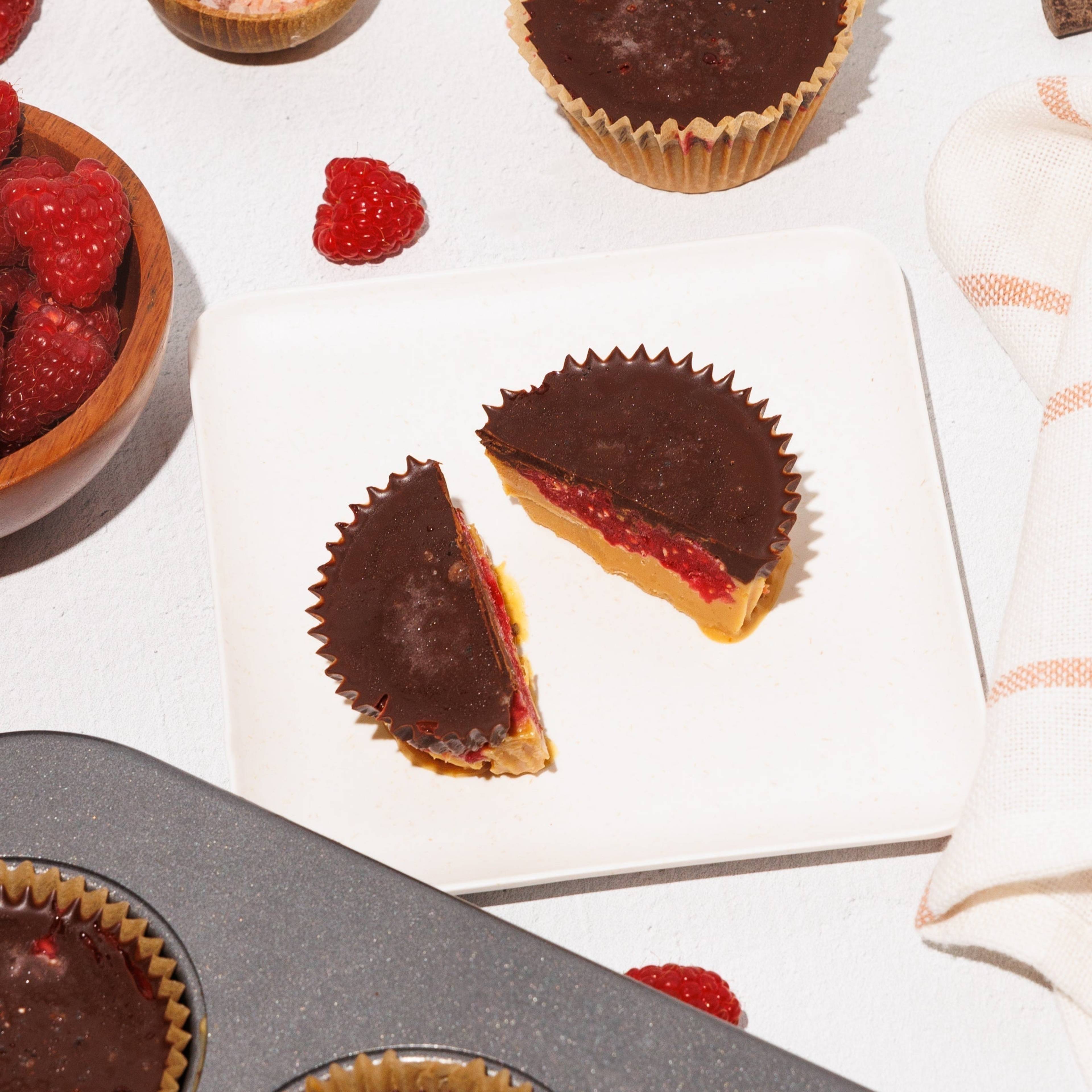 Chocolate-topped cups, including a cut-open cup with red filling and a yellow base, alongside raspberries, chocolate chunks, and pink salt.