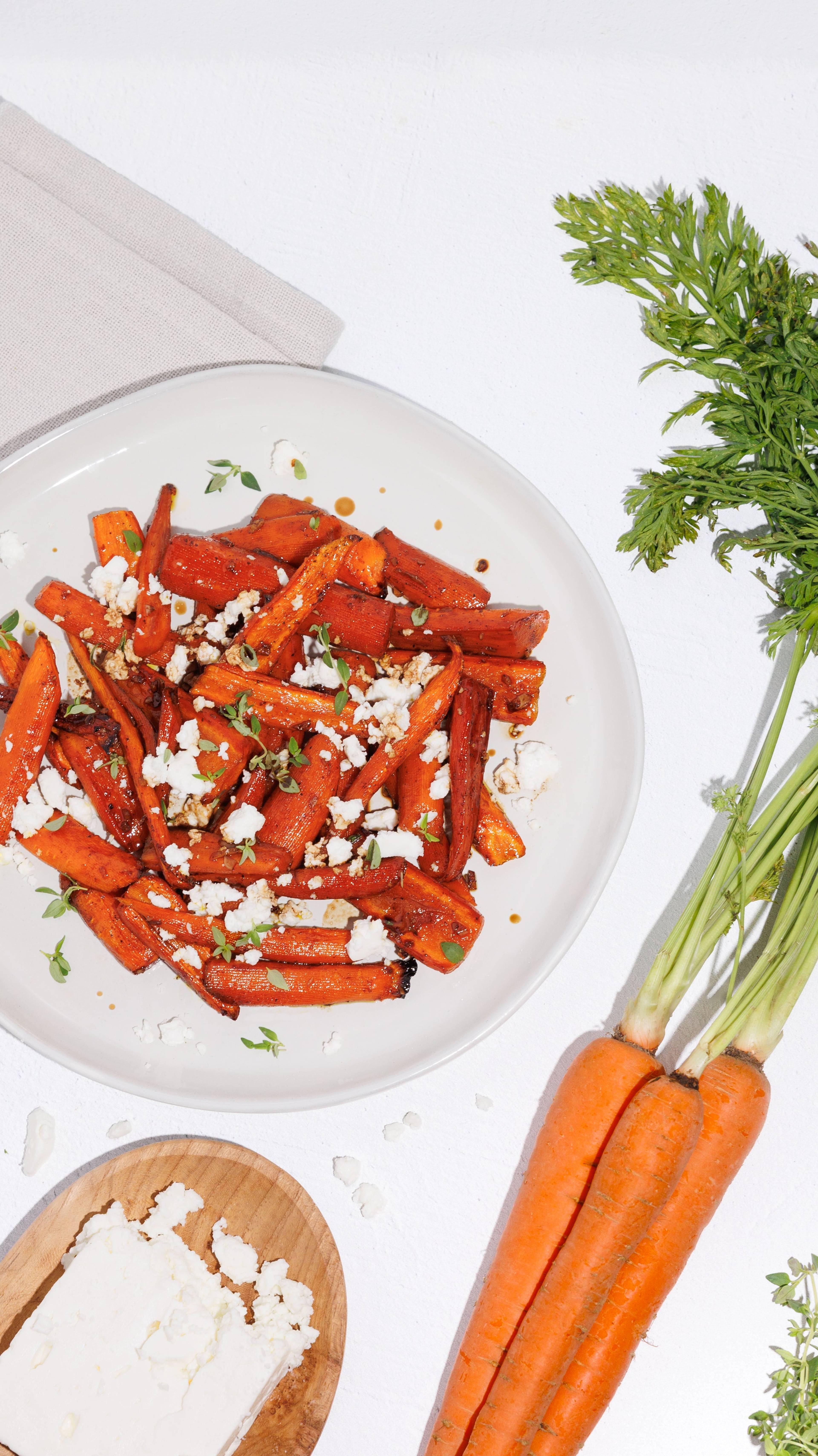Roasted carrots with crumbled cheese and herbs on a white plate beside fresh carrots.