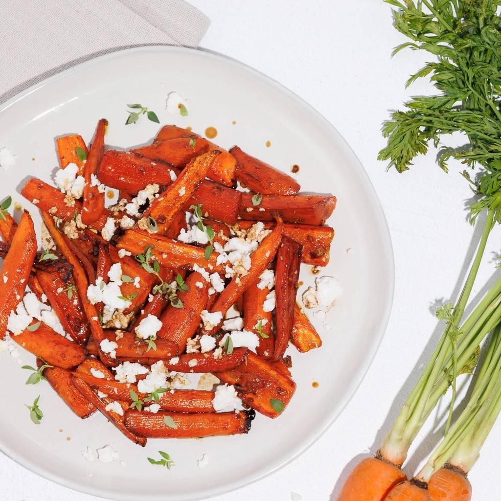 Roasted carrots with crumbled cheese and herbs on a white plate beside fresh carrots.