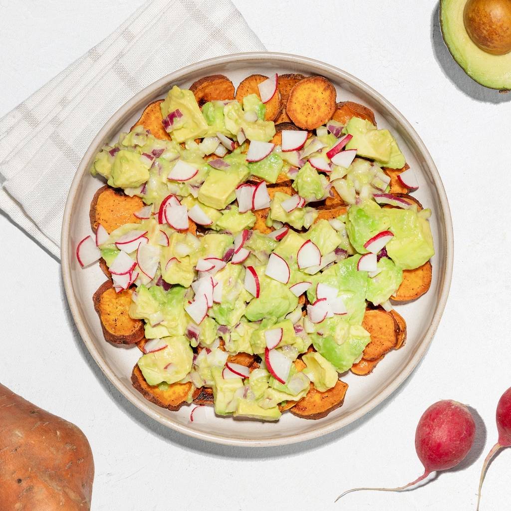 Plate of sweet potato rounds topped with avocado, radish, and diced onion, with an avocado half and whole vegetables nearby.