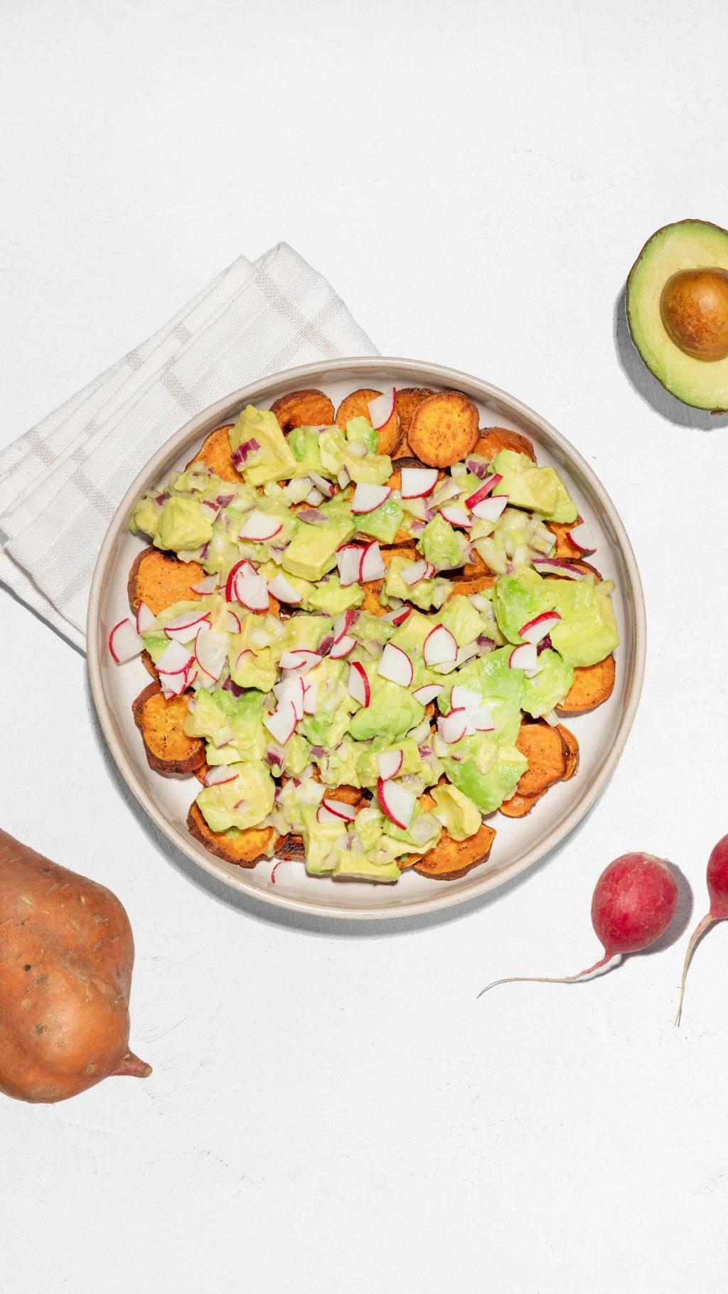 Plate of sweet potato rounds topped with avocado, radish, and diced onion, with an avocado half and whole vegetables nearby.