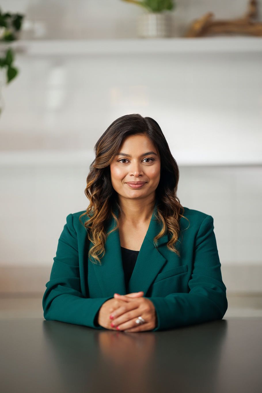 Woman with dark wavy hair in a teal blazer sits at a table with her hands folded.