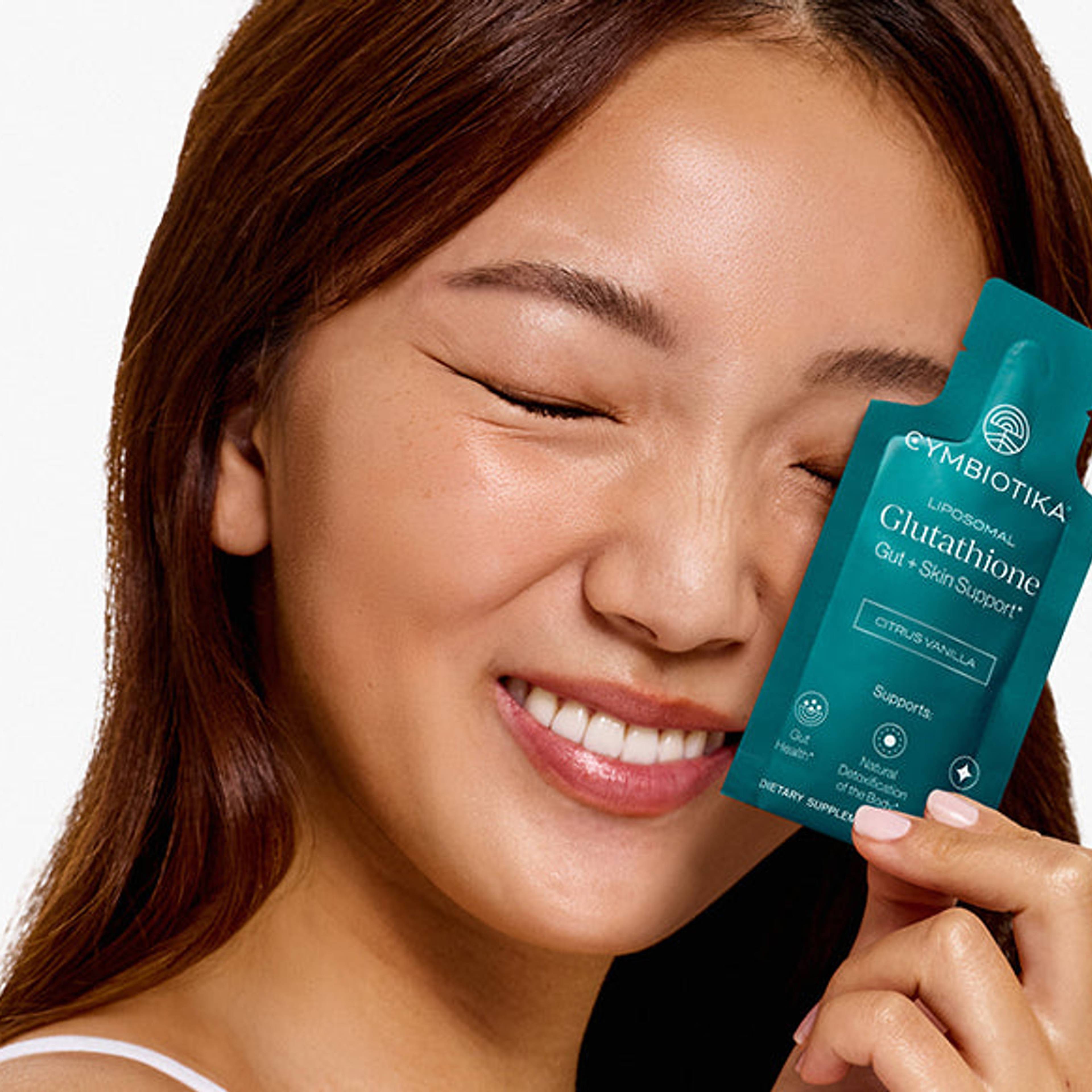 Smiling woman holding a Cymbiotika Liposomal Glutathione packet beside her face.