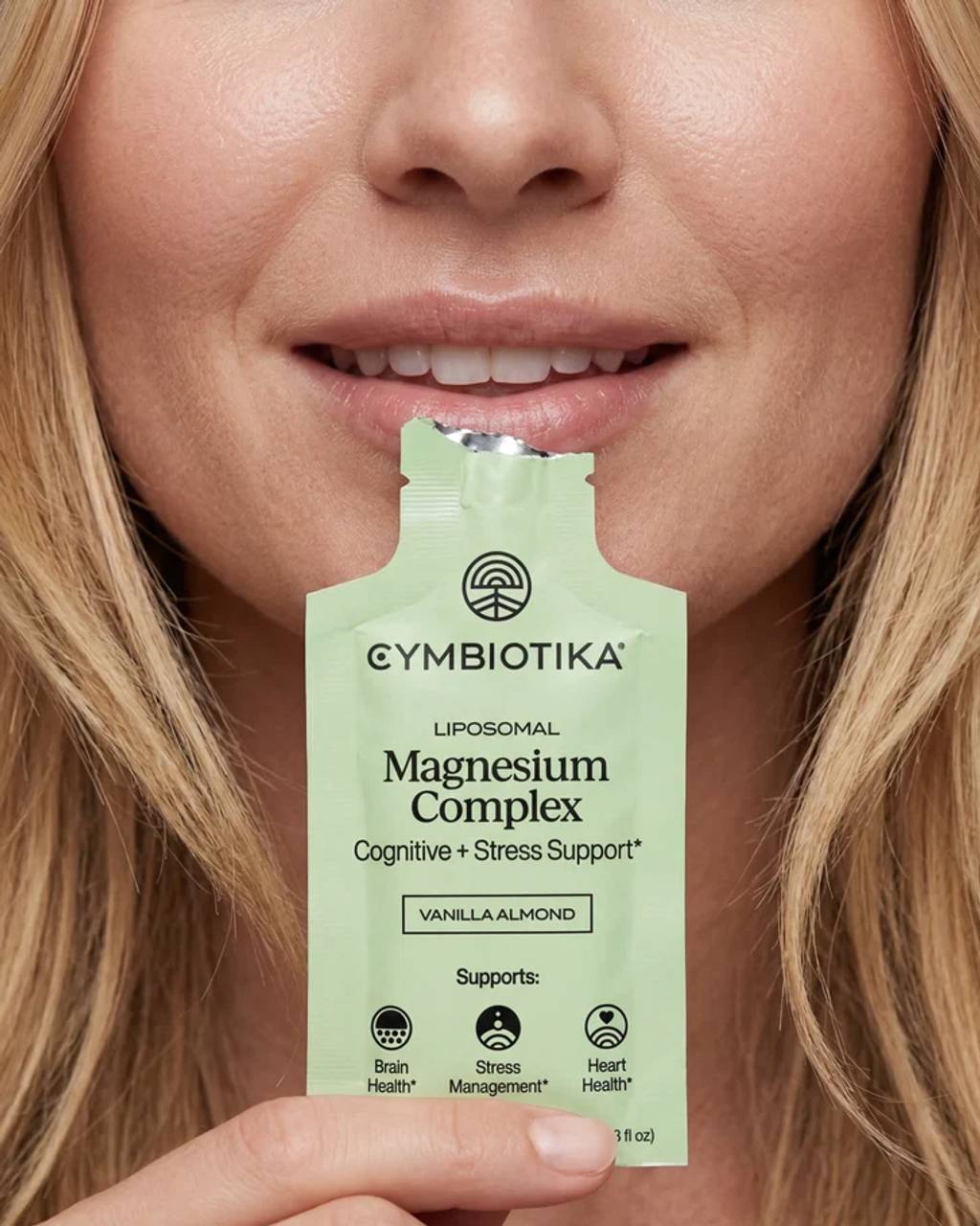 A woman opens a Liposomal Magnesium Complex sachet with her mouth; the pale green packet is held in her hand.