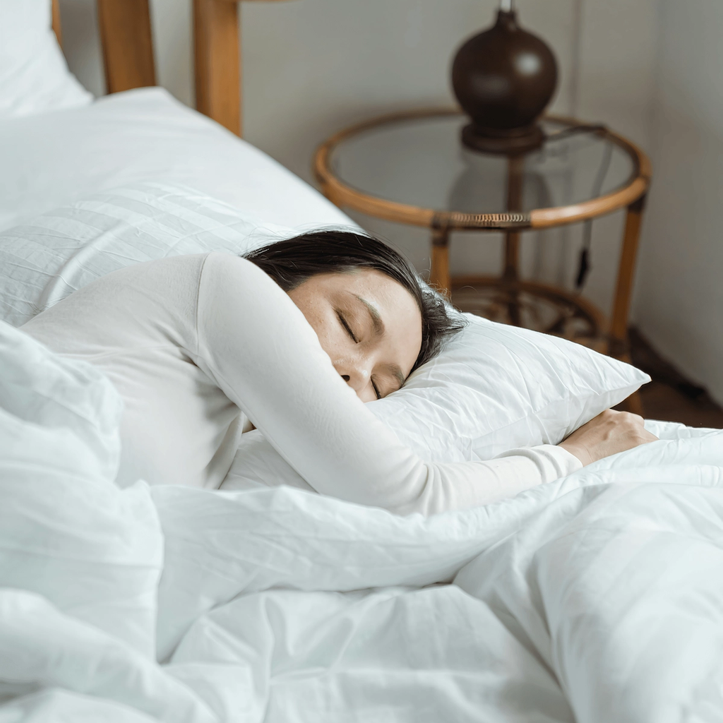 Woman sleeping on her side in a white bed beside a bedside lamp.