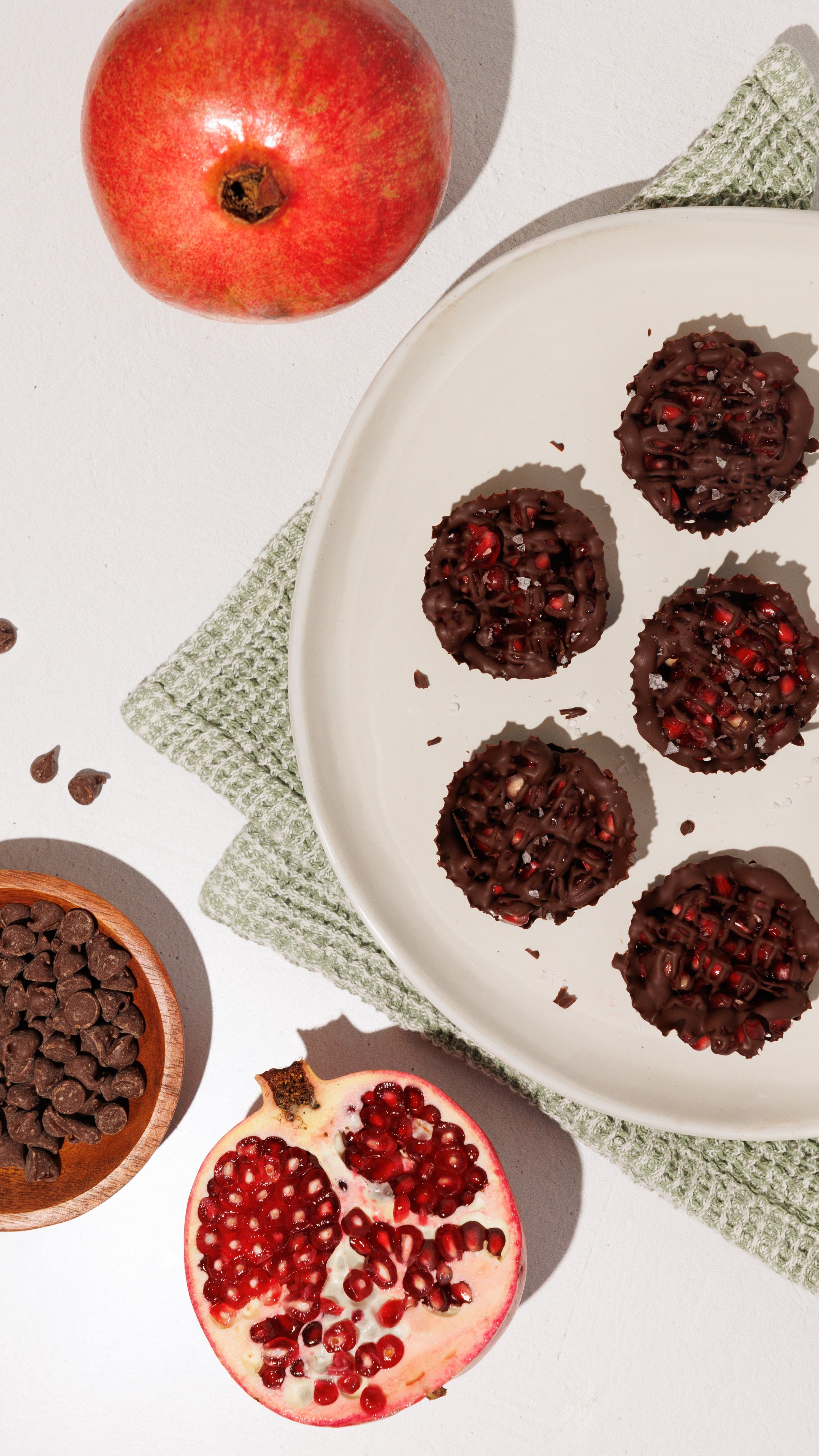 Chocolate pomegranate cups on a white plate with whole and halved pomegranates and chocolate chips.