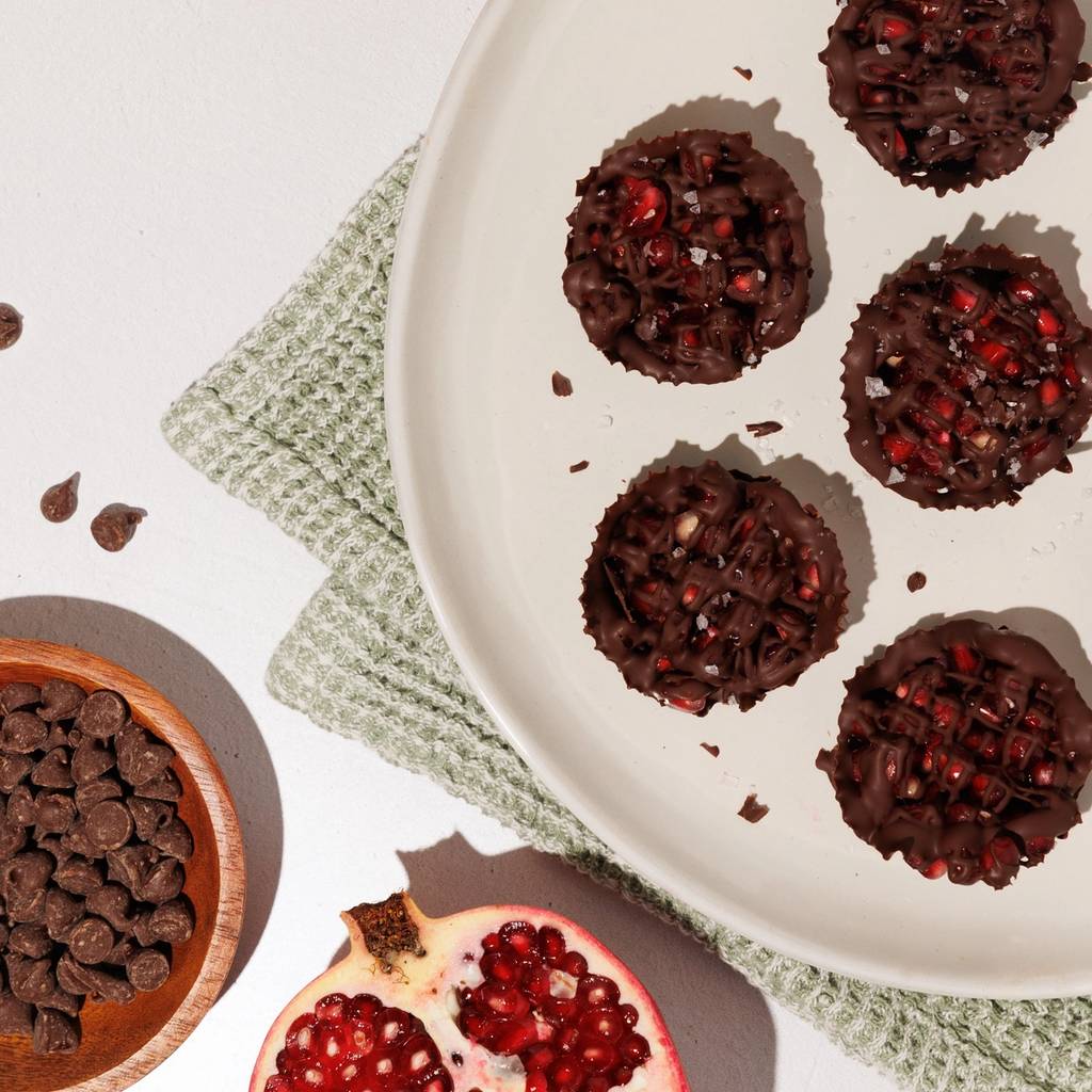 Chocolate pomegranate cups on a white plate with whole and halved pomegranates and chocolate chips.