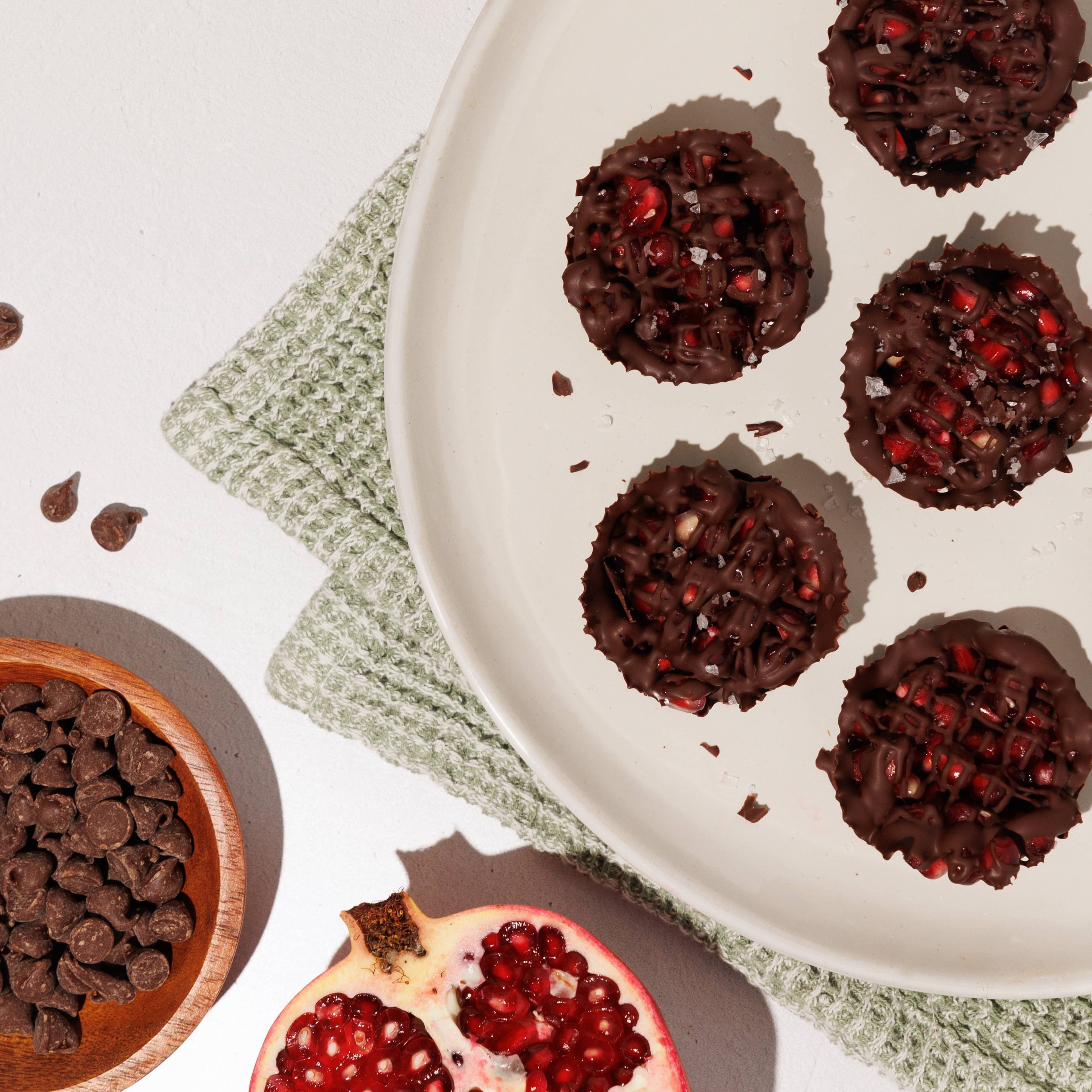 Chocolate pomegranate cups on a white plate with whole and halved pomegranates and chocolate chips.