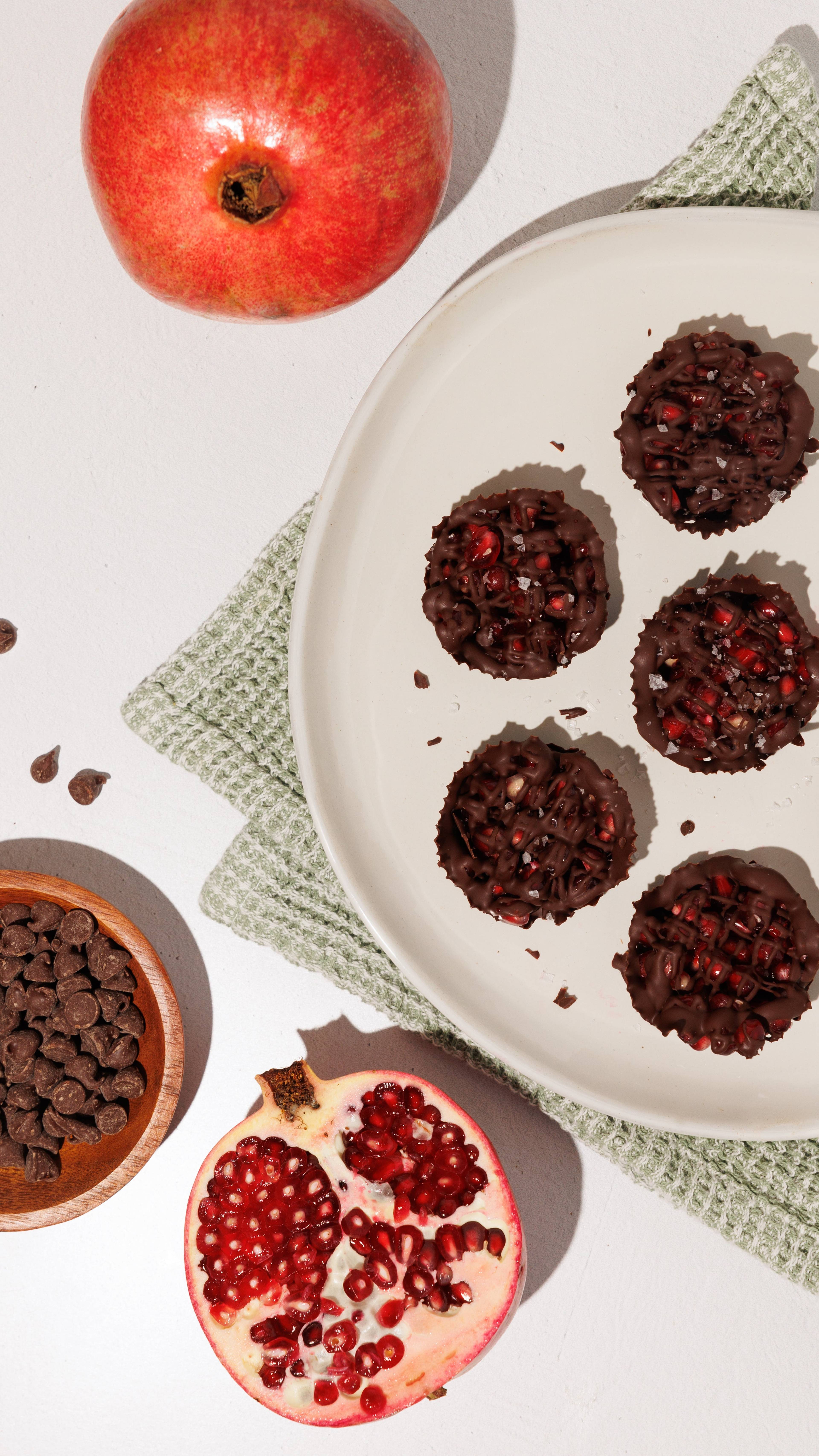 Chocolate pomegranate cups on a white plate with whole and halved pomegranates and chocolate chips.