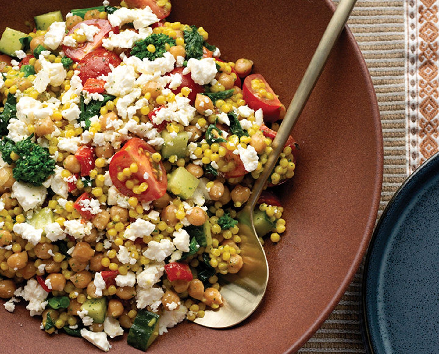 Couscous salad with chickpeas, cherry tomatoes, cucumber, greens, corn, and crumbled feta on a brown plate.