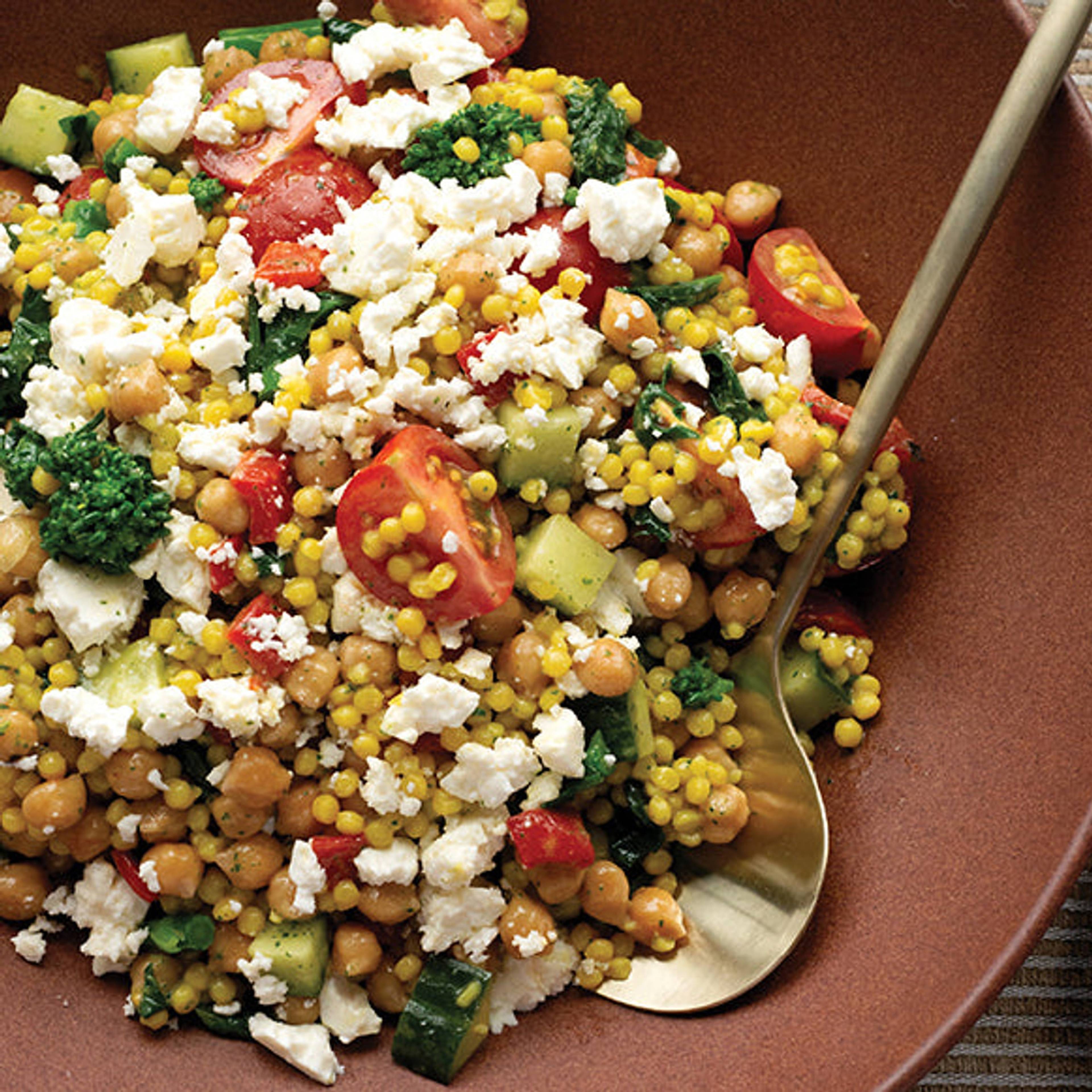 Couscous salad with chickpeas, cherry tomatoes, cucumber, greens, corn, and crumbled feta on a brown plate.