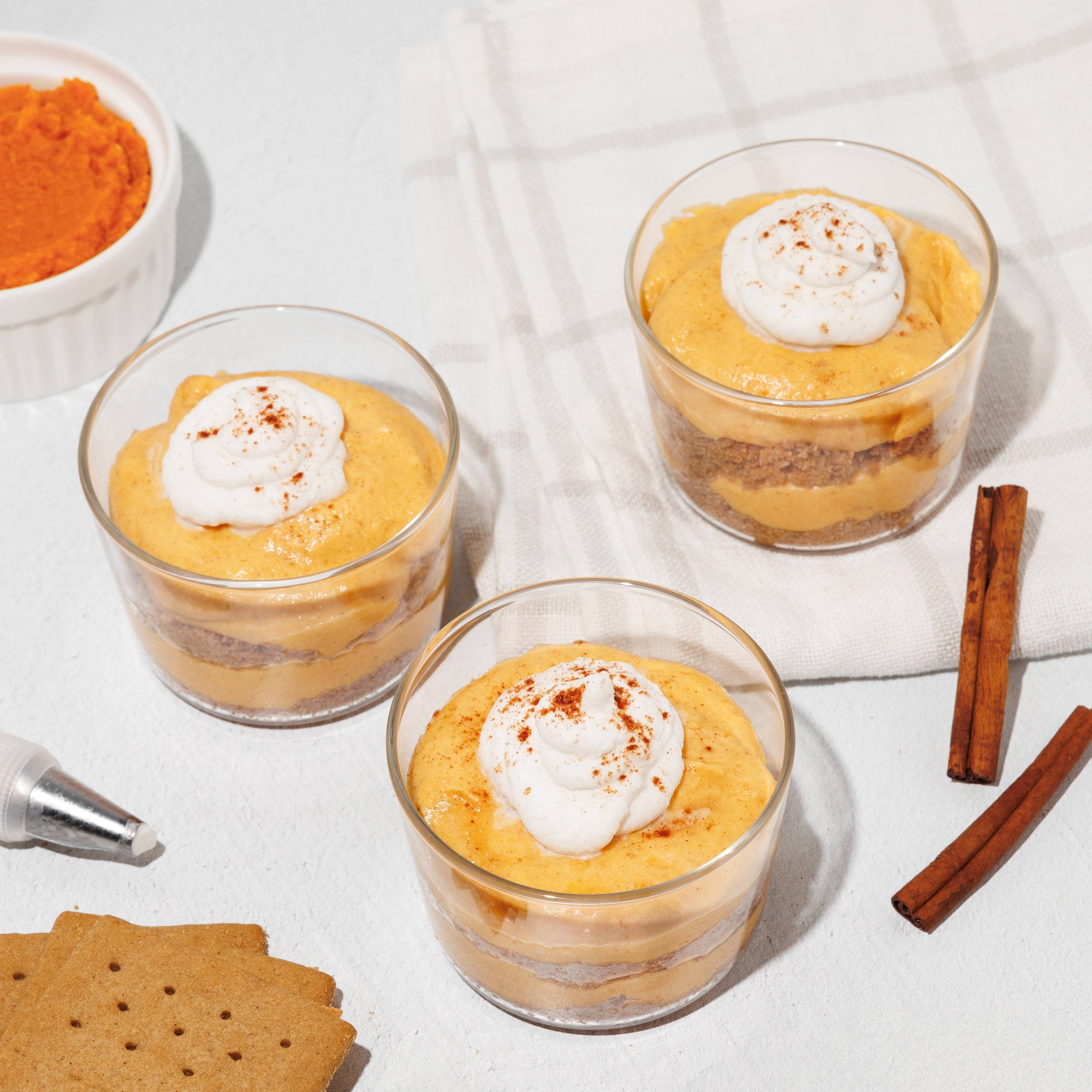 Three layered orange desserts topped with whipped cream, with crackers, cinnamon sticks, and bowls of ingredients nearby.